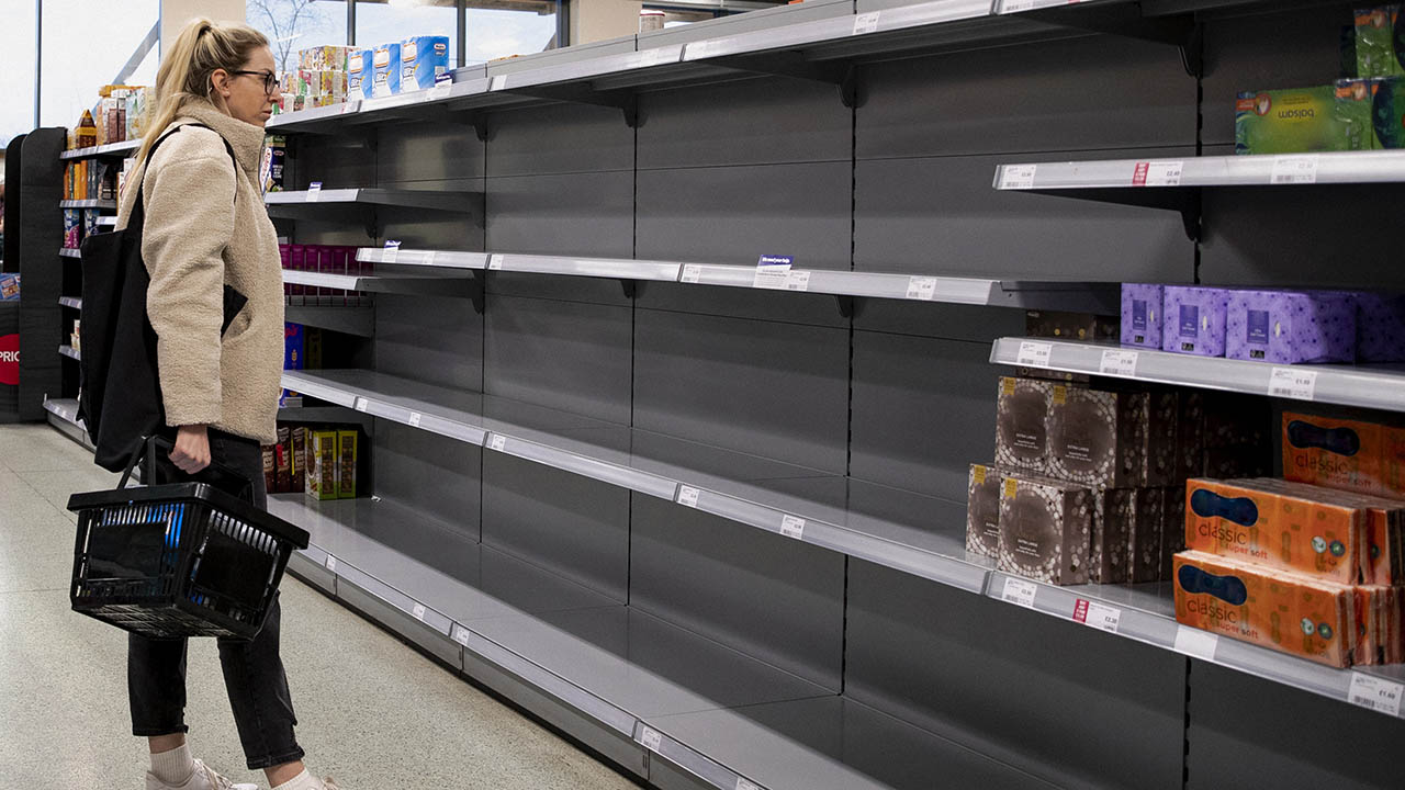 A young Caucasian female stands looking concerned at the empty shelves in a supermarket.