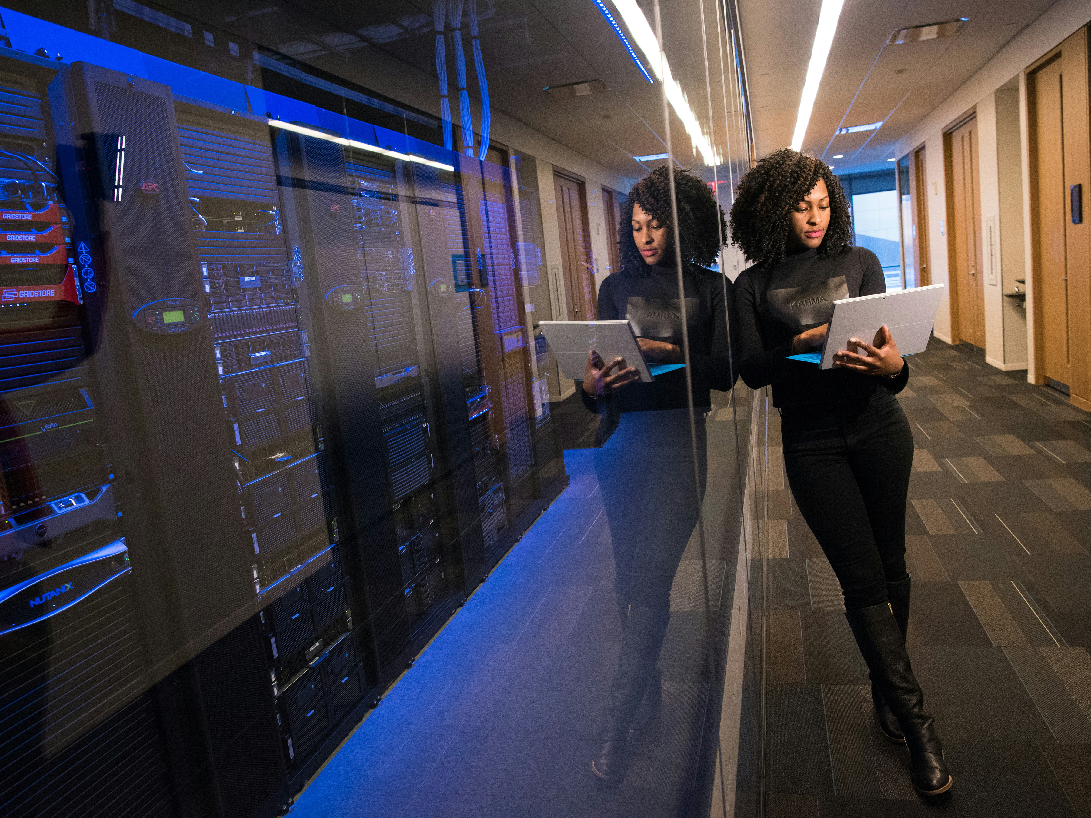A black engineer stands outside a server room looking at her laptop screen