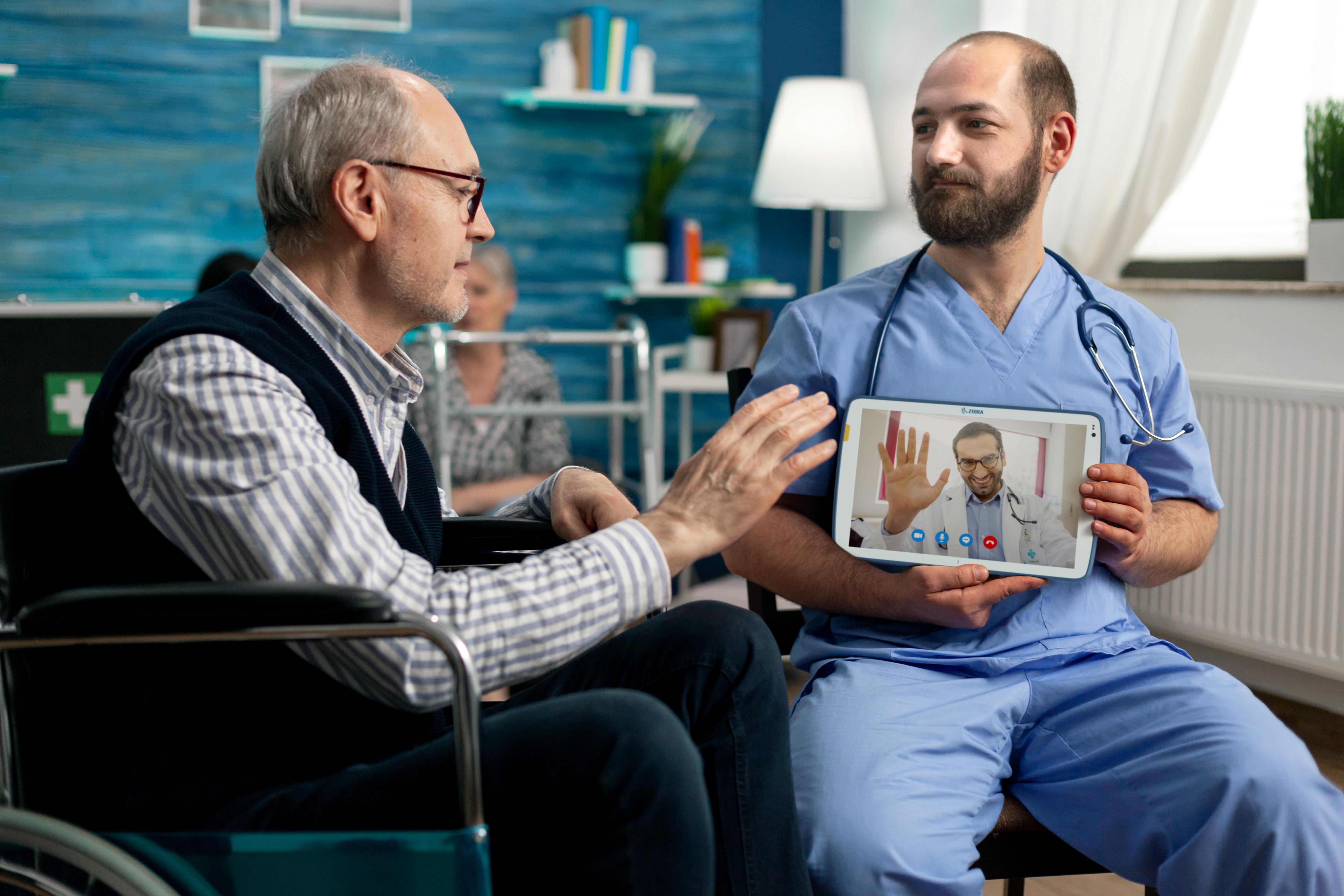 Retired disabled man video calling with his family in nursing home while health care assistant holds electronic device to assist. Wheelchair patient talking to daughter and granddaughter remotely.