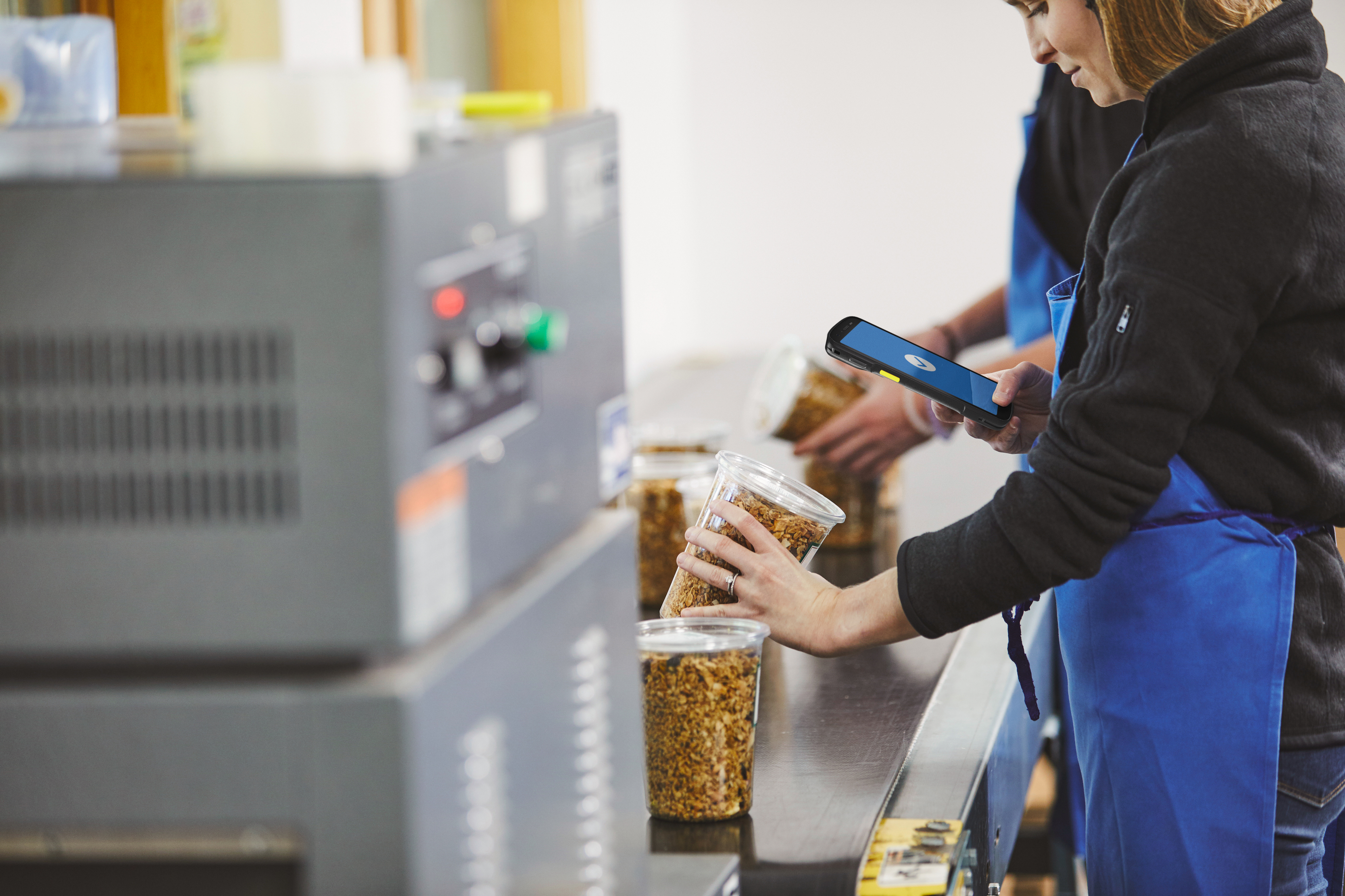 A worker using a handheld mobile computer to scan food products on a conveyor belt inside of a manufacturing plant.