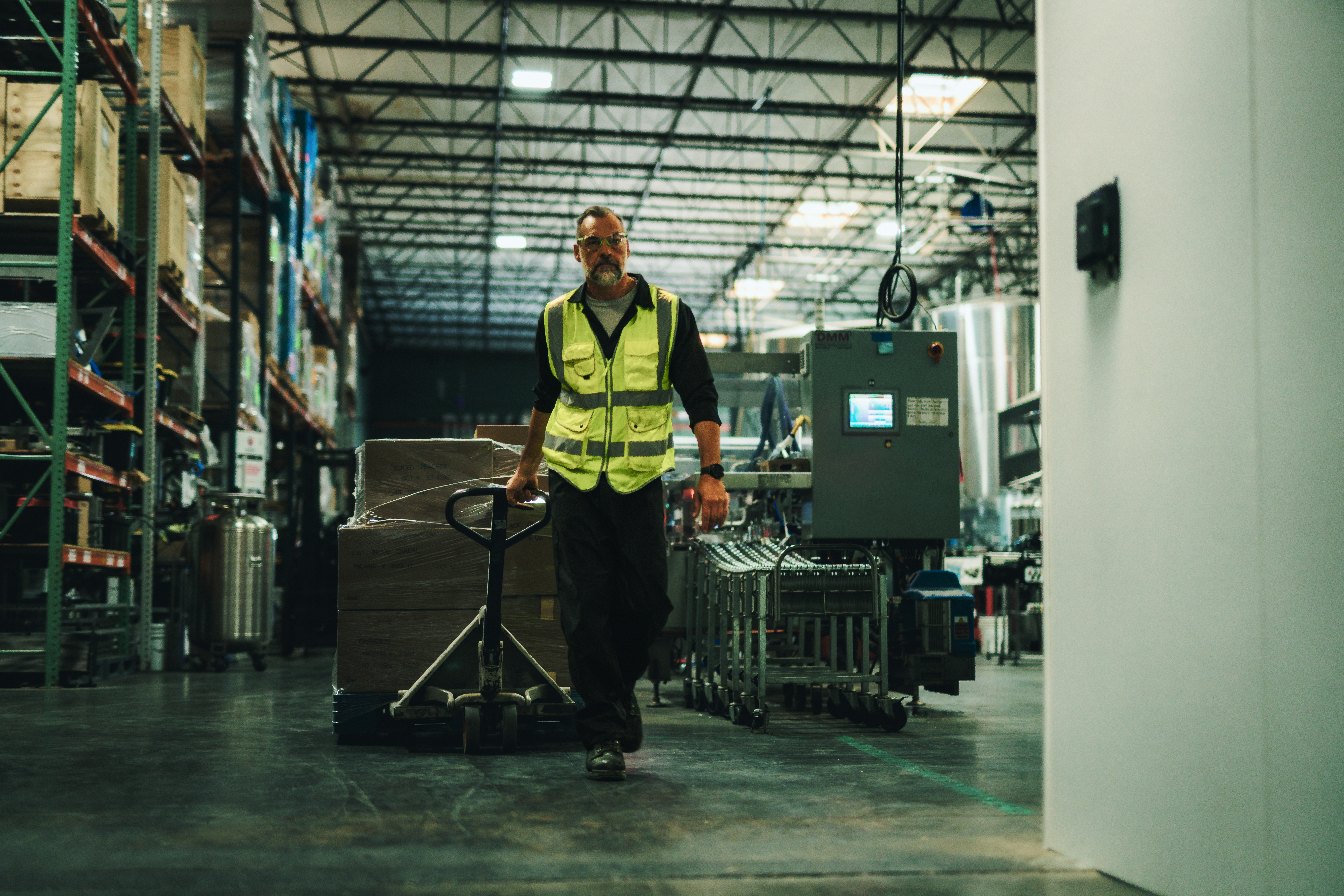 Warehouse worker in a safety vest moves a loaded pallet through a manufacturing facility past a black Zebra FXR60 fixed RFID reader.