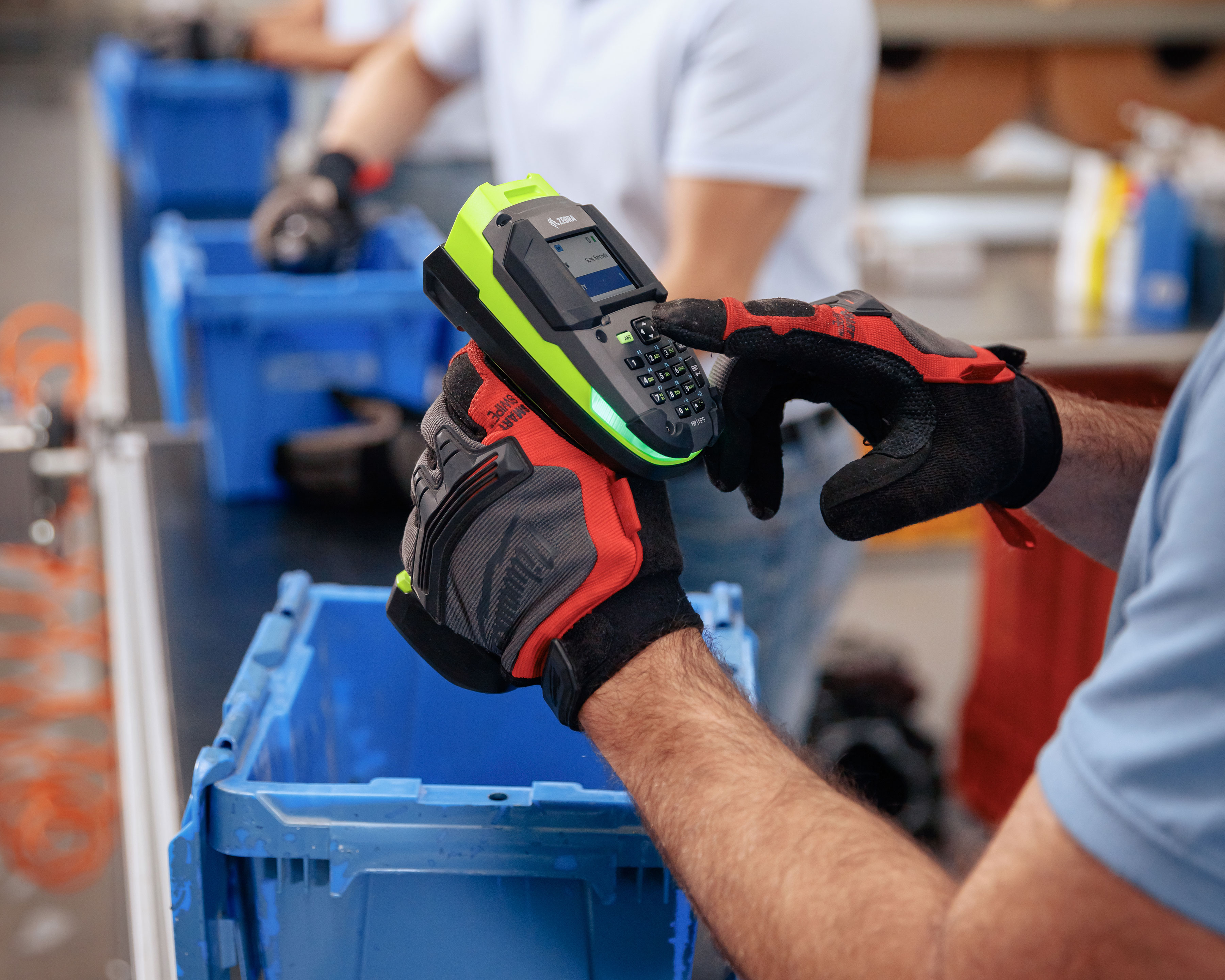 Worker uses the keypad of Zebra DS3600 barcode scanner in a manufacturing plant as other workers pack products in blue storage bins