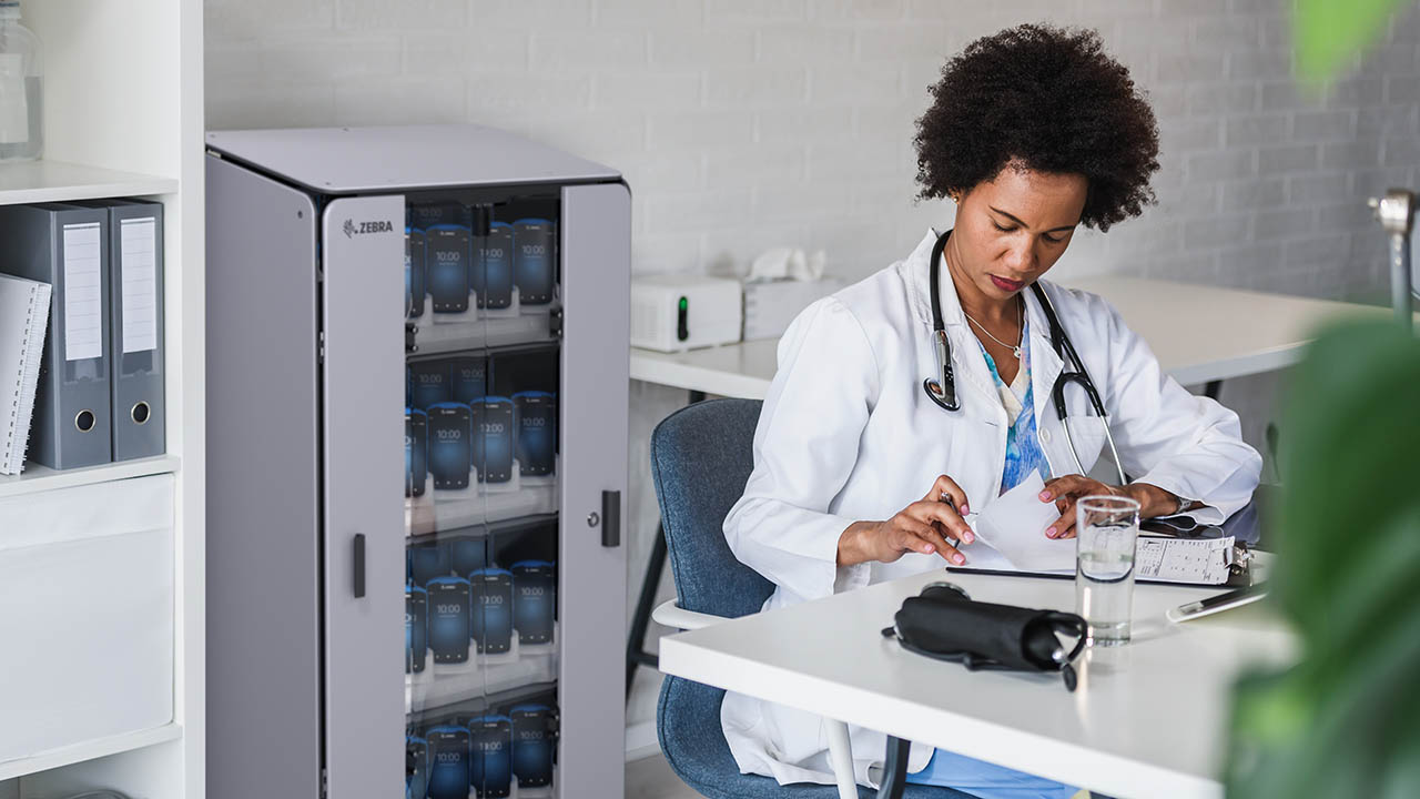 A doctor sits in an office with Zebra healthcare mobile computers charging behind her in an intelligent cabinet. 