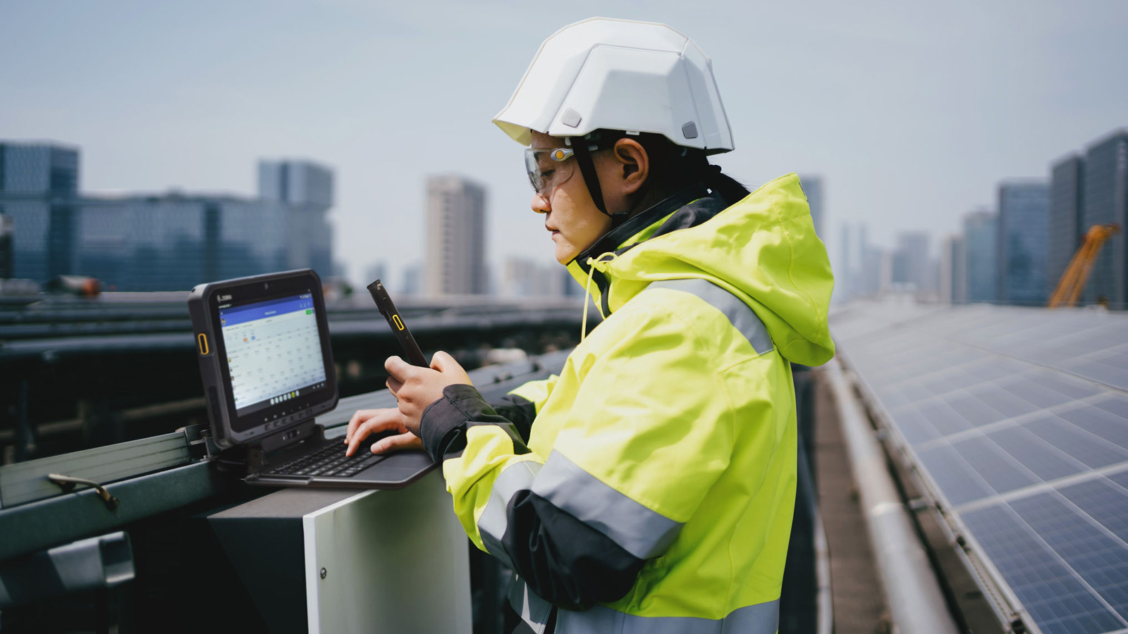 A field technician works on a rooftop solar farm and is operating a rugged Zebra Tablet mounted on a keyboard base while holding a Zebra Handheld mobile computer