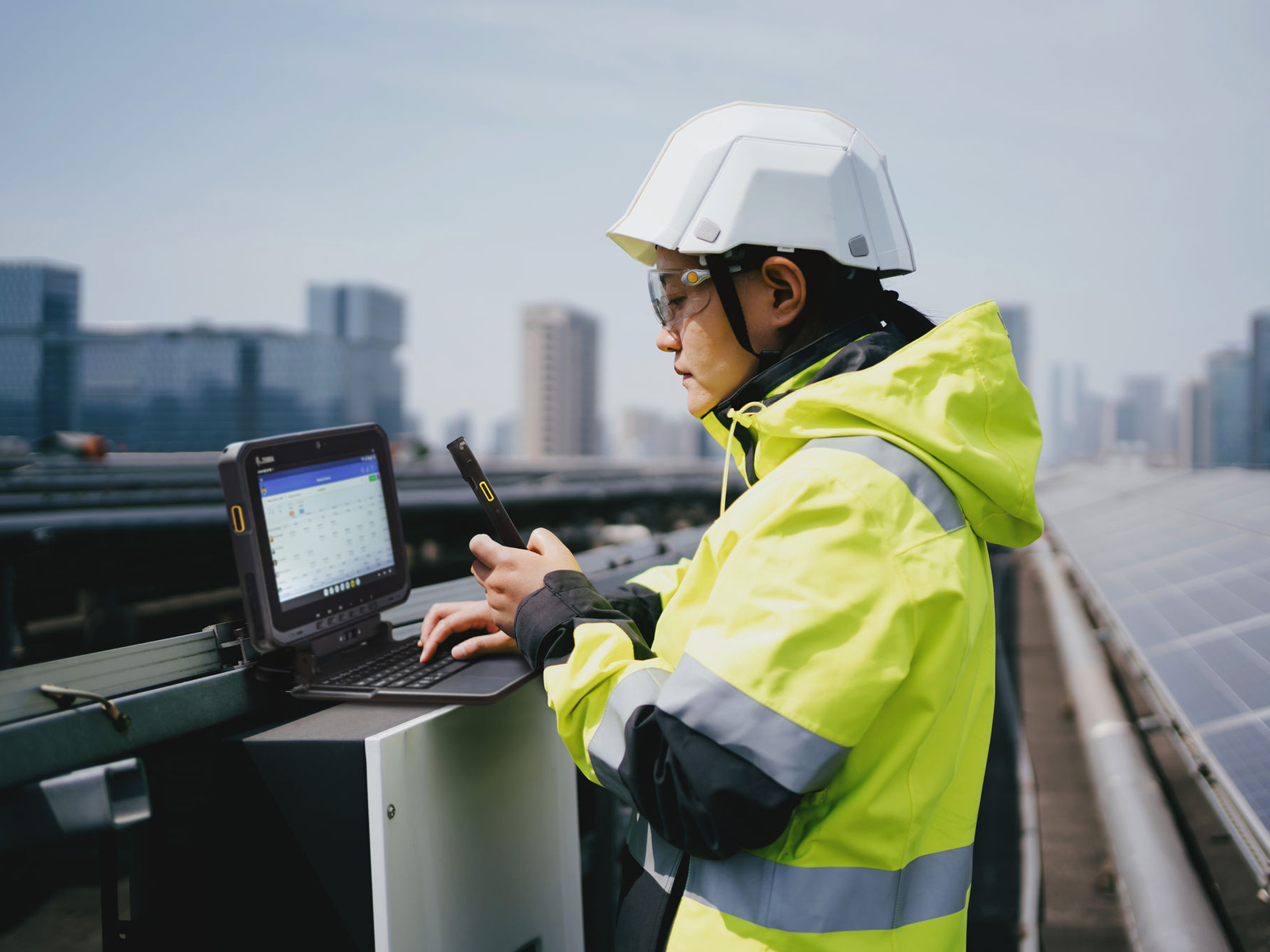 A field technician works on a rooftop solar farm and is operating a rugged Zebra Tablet mounted on a keyboard base while holding a Zebra Handheld mobile computer