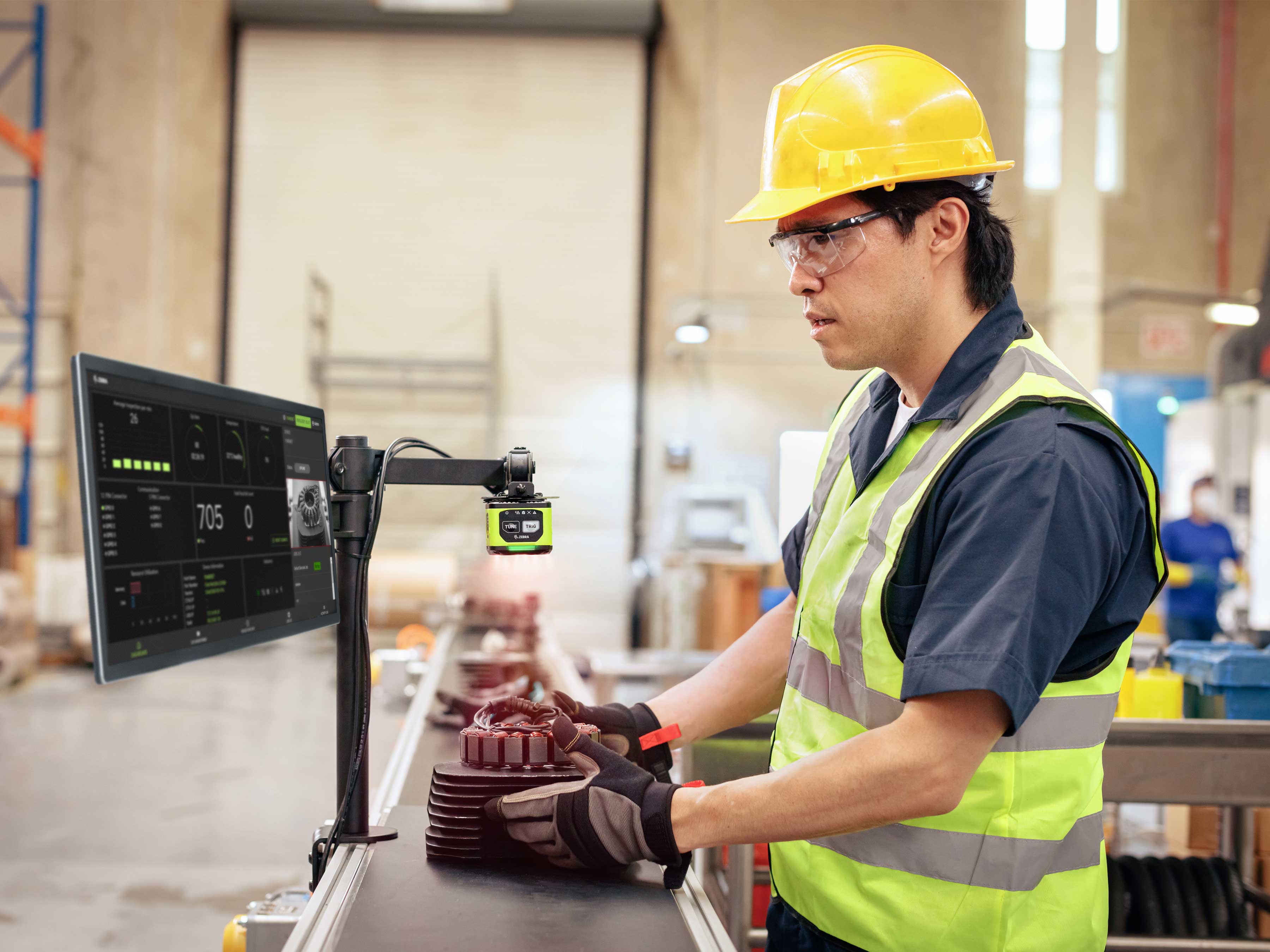 A manufacturing worker performs a quality check on a part using a Zebra FS40 Fixed Industrial Scanner and VS40 Smart Camera, with real-time analytics displayed on a monitor.