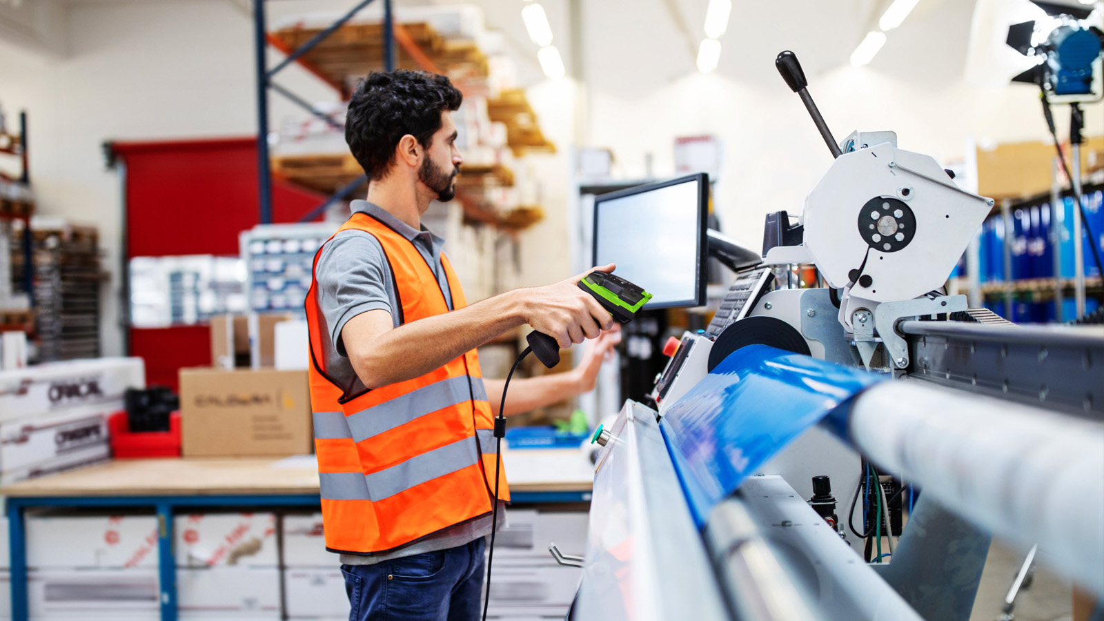 A factory worker in an orange safety vest uses a Zebra 3600 Series Ultra-Rugged handheld barcode scanner in a busy manufacturing environment.