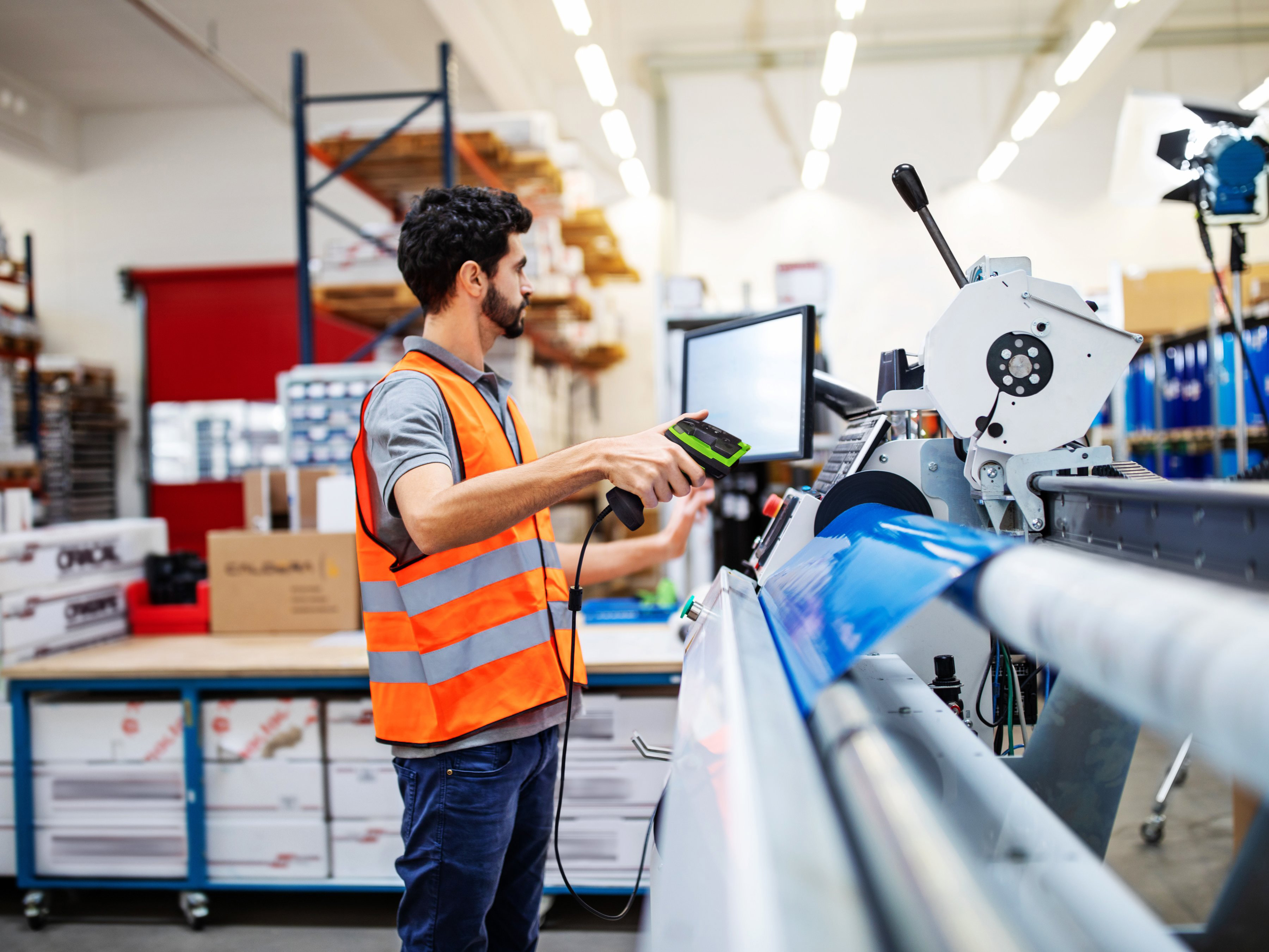 A factory worker in an orange safety vest uses a Zebra 3600 Series Ultra-Rugged handheld barcode scanner in a busy manufacturing environment.