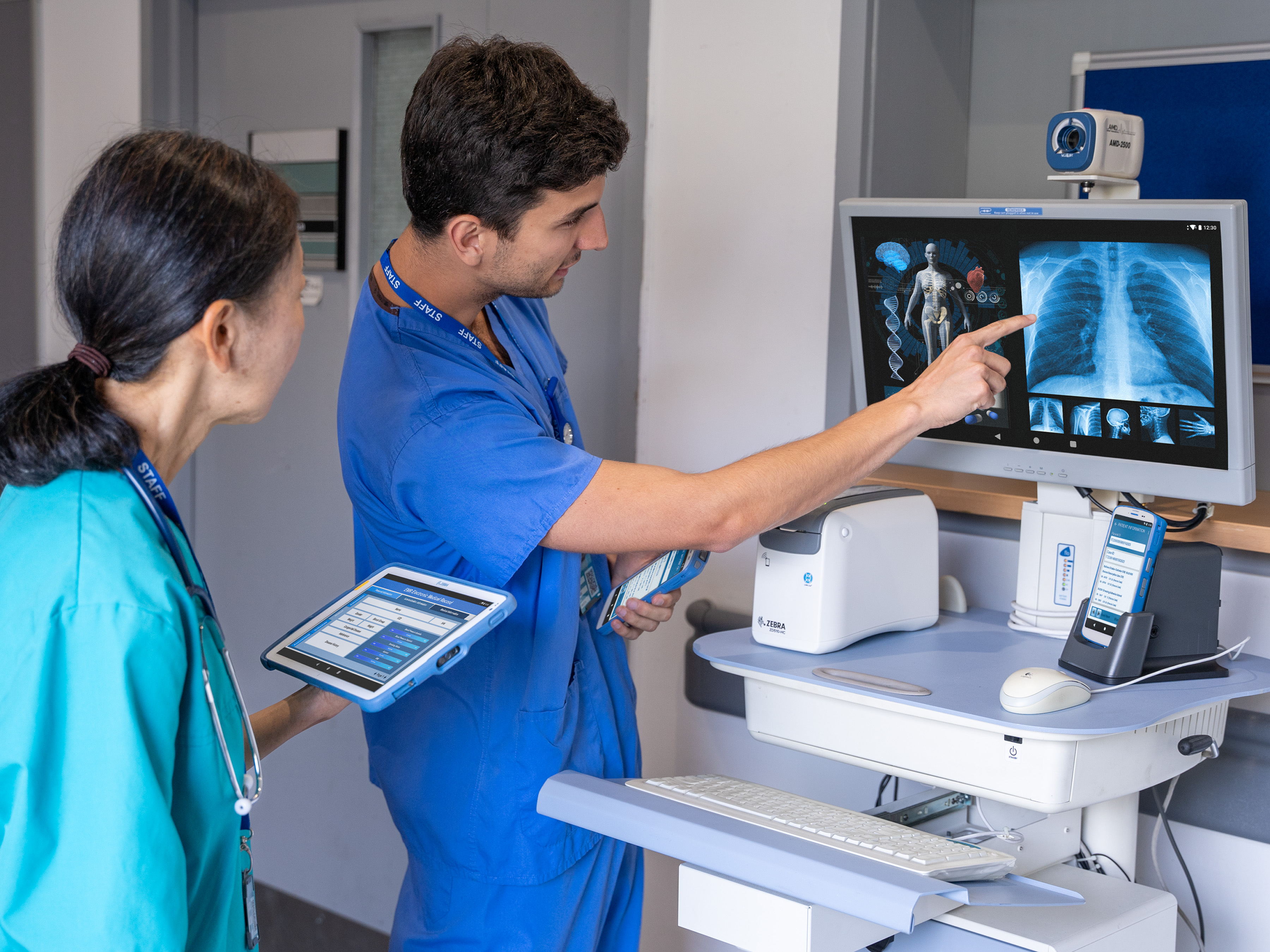 What Is an Electronic Health Record (EHR) Two healthcare practitioners collaborate in a hospital room setting. A woman, holding a tablet, looks at a computer screen as a man points to the X-ray displayed, explaining it to her.
