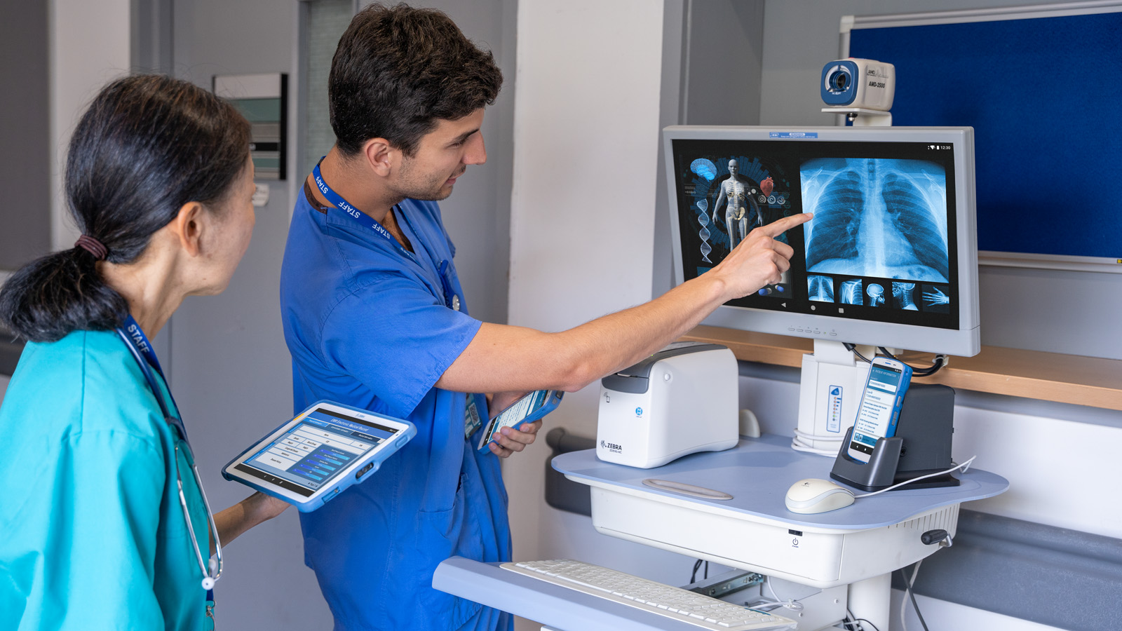 What Is an Electronic Health Record (EHR) Two healthcare practitioners collaborate in a hospital room setting. A woman, holding a tablet, looks at a computer screen as a man points to the X-ray displayed, explaining it to her.