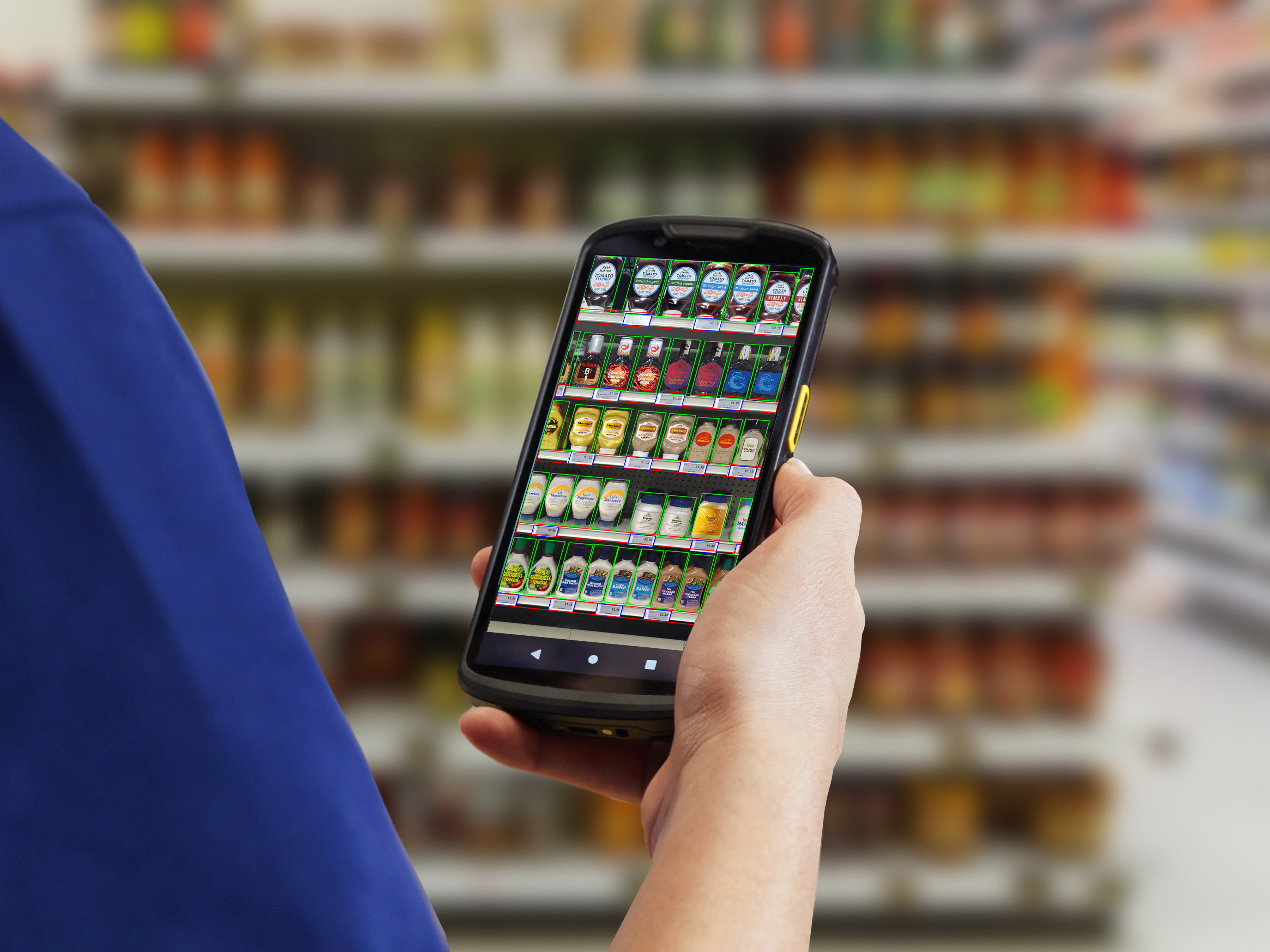 An employee in a market is looking through a Zebra mobile computer, using the Zebra mobile computing AI suite to locate a product on a shelf.