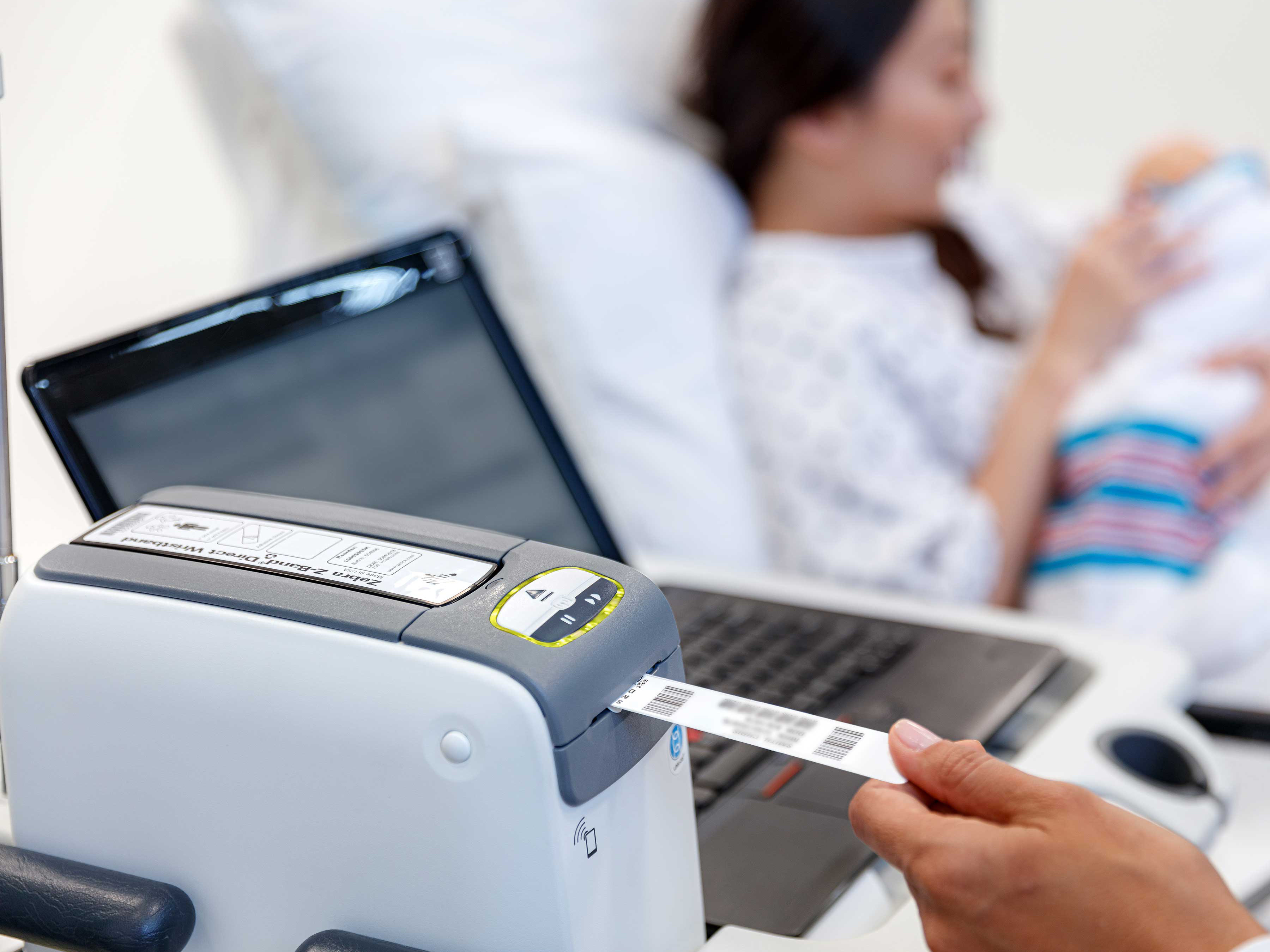 The Zebra Patient Identification wristband is being printed using a Zebra printer next to a patient holding a newborn while lying on a bed. 