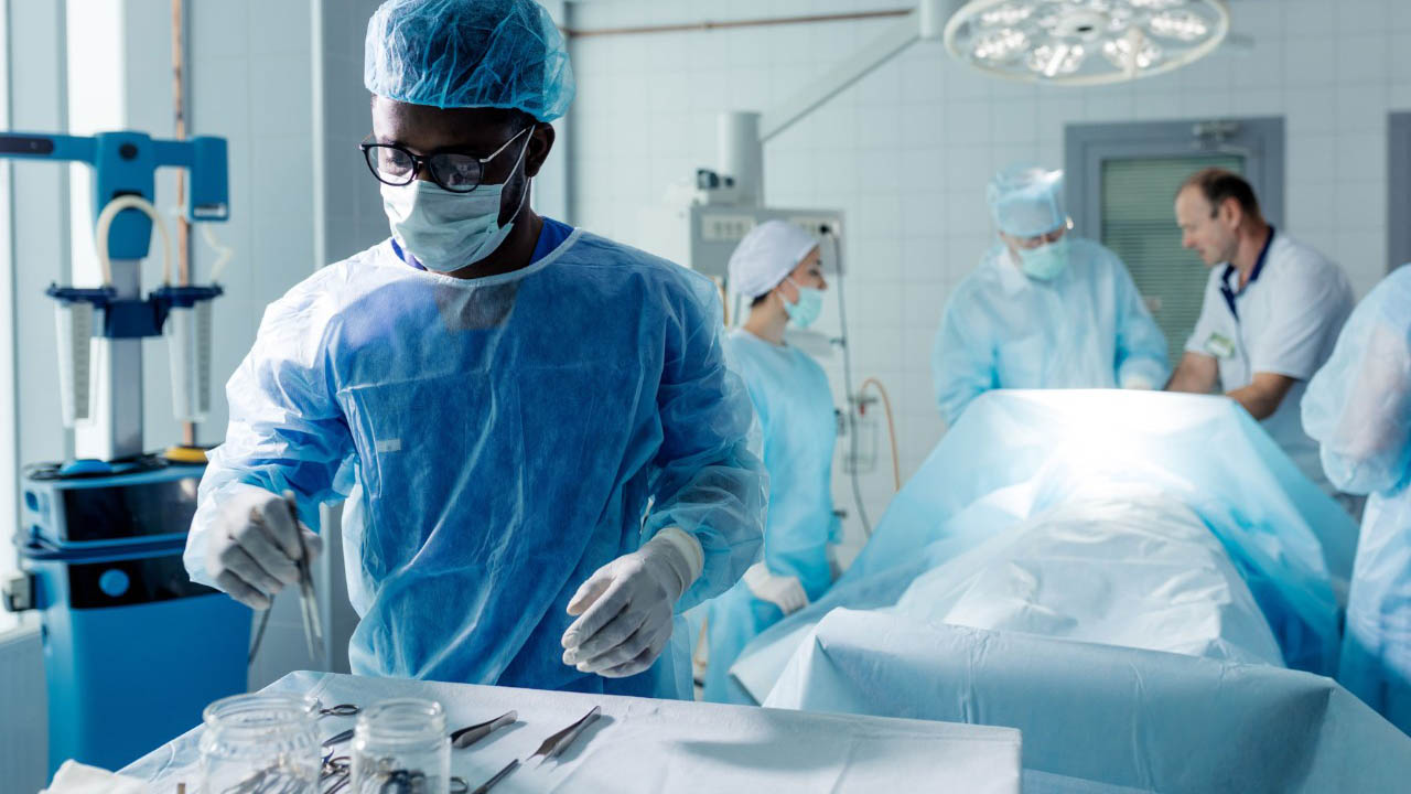 An operating room team member reaches for sterile instruments off the tray while surgeons work in the background