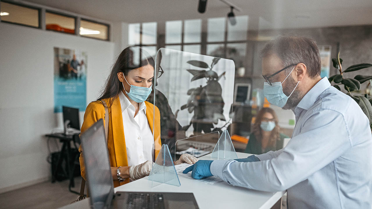 A bank teller assists a customer from behind glass while both wear masks