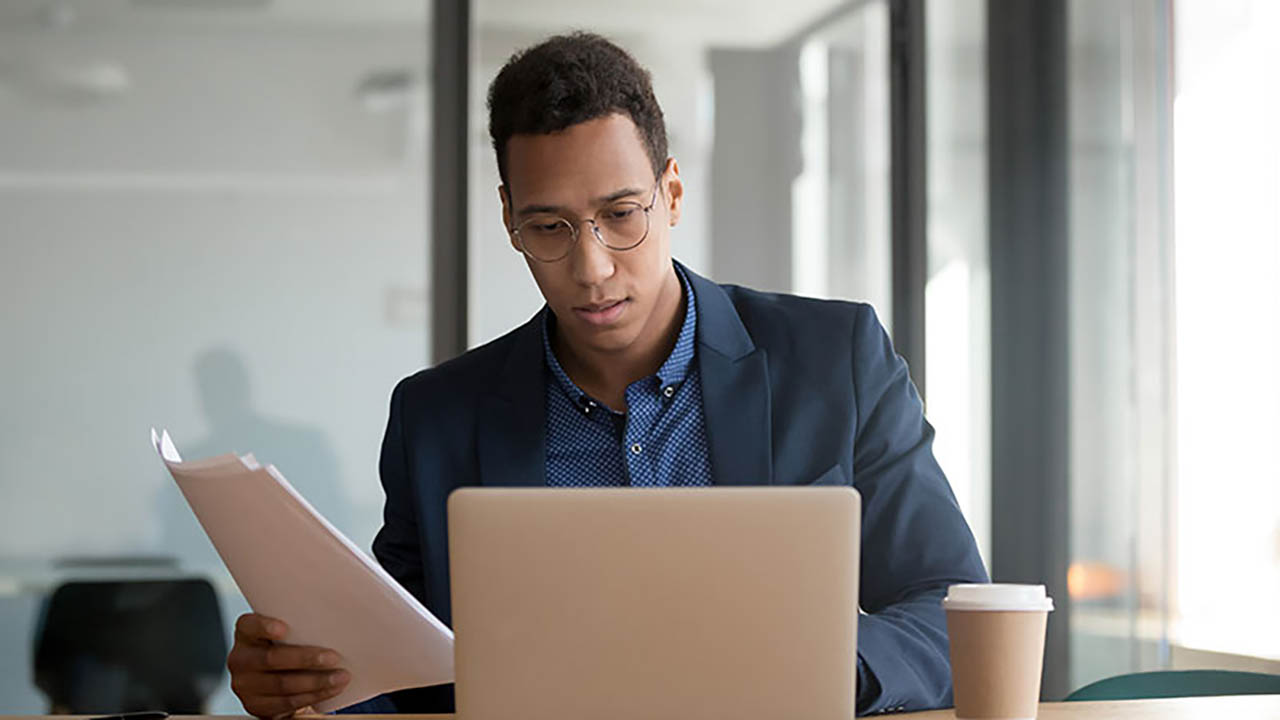 A banking associate reviews paperwork and tasks on his laptop screen