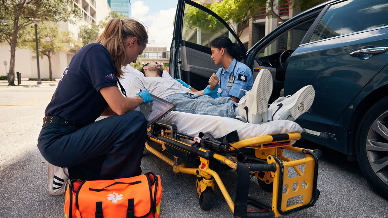 Two EMTs attend to a patient near a car. One inputs data into a rugged tablet.