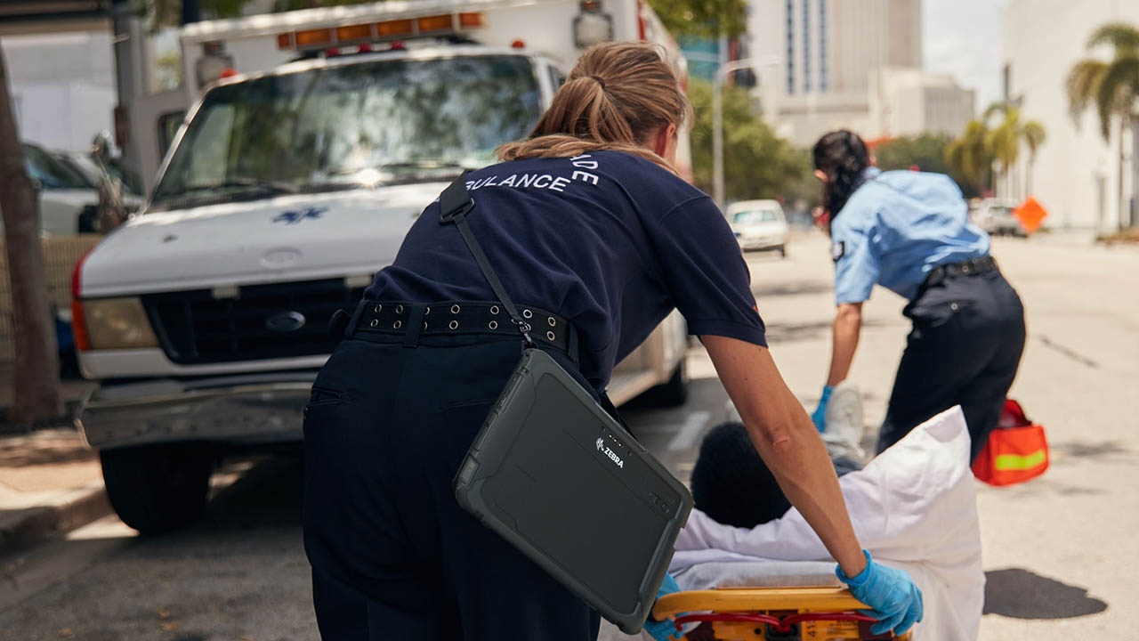 A paramedic carries a rugged tablet on her shoulder while moving a patient on a gurney