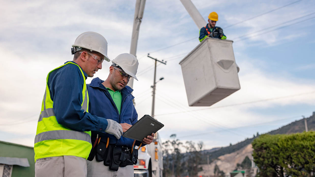 Two utility workers look at an ET85 rugged tablet on the ground while another crew member in a bucket services a power pole