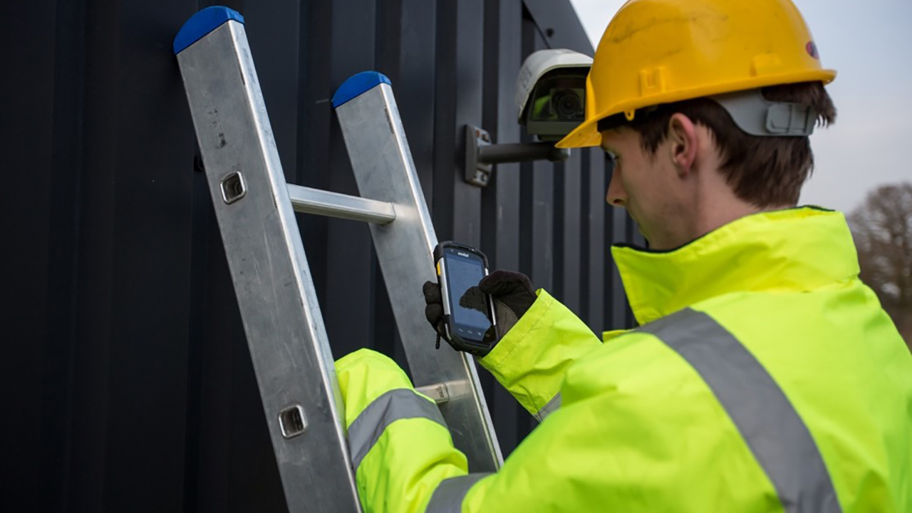 Male field technician climbs a ladder on the side of a building with a rugged mobile computer in hand