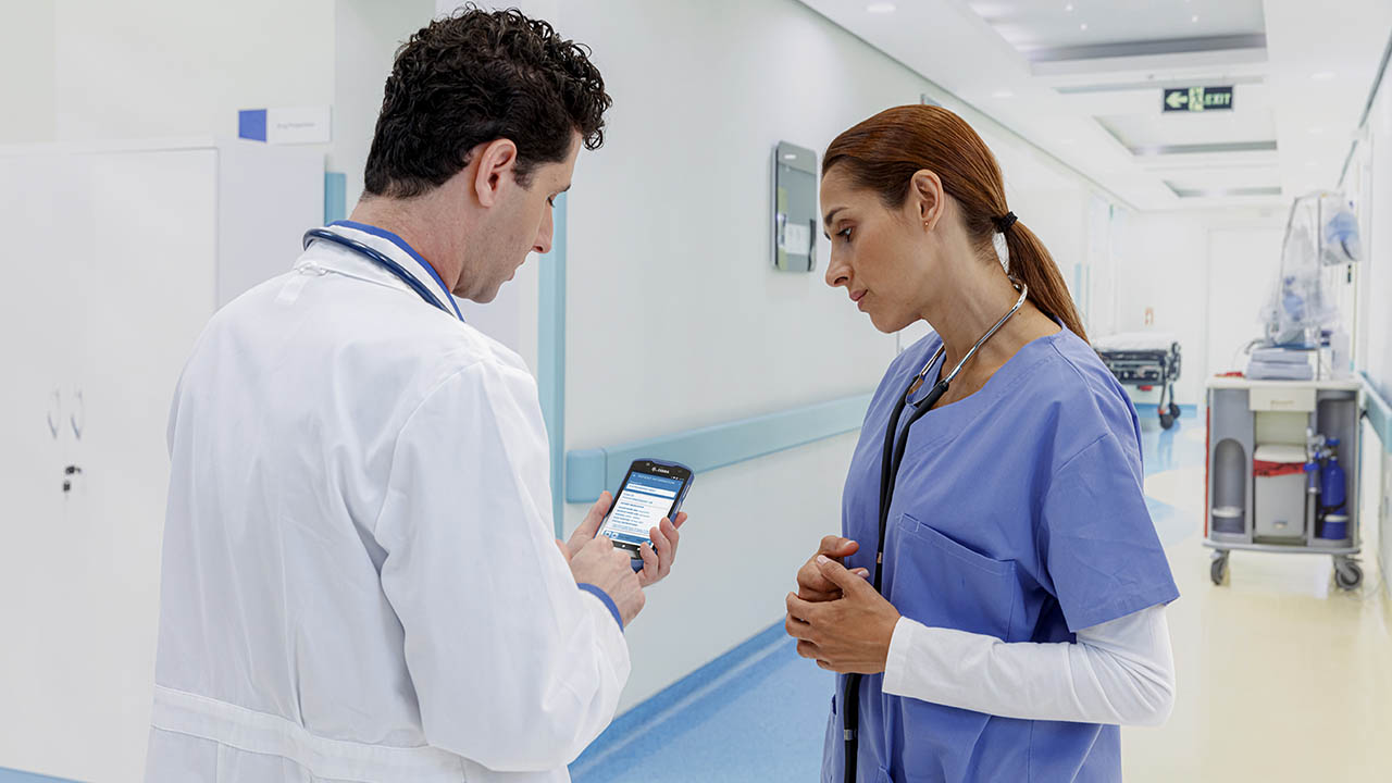A doctor and nurse speak in the hallway while the doctor looks at his clinical smartphone