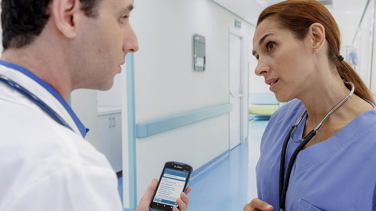 A doctor and nurse speak in the hallway while the doctor looks at his Zebra TC52ax-HC clinical smartphone