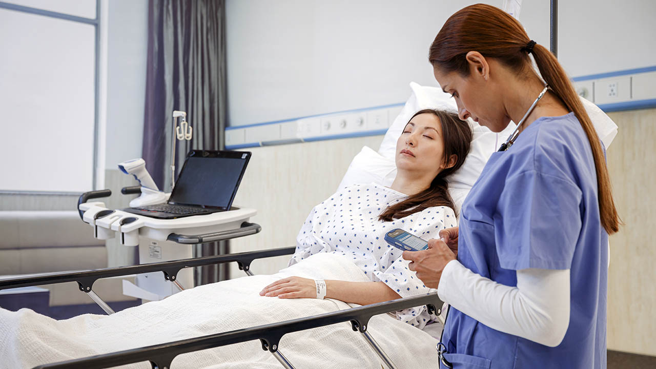 A nurse looks at her Zebra TC52ax-HC clinical smartphone while standing at a sleeping patient's bedside