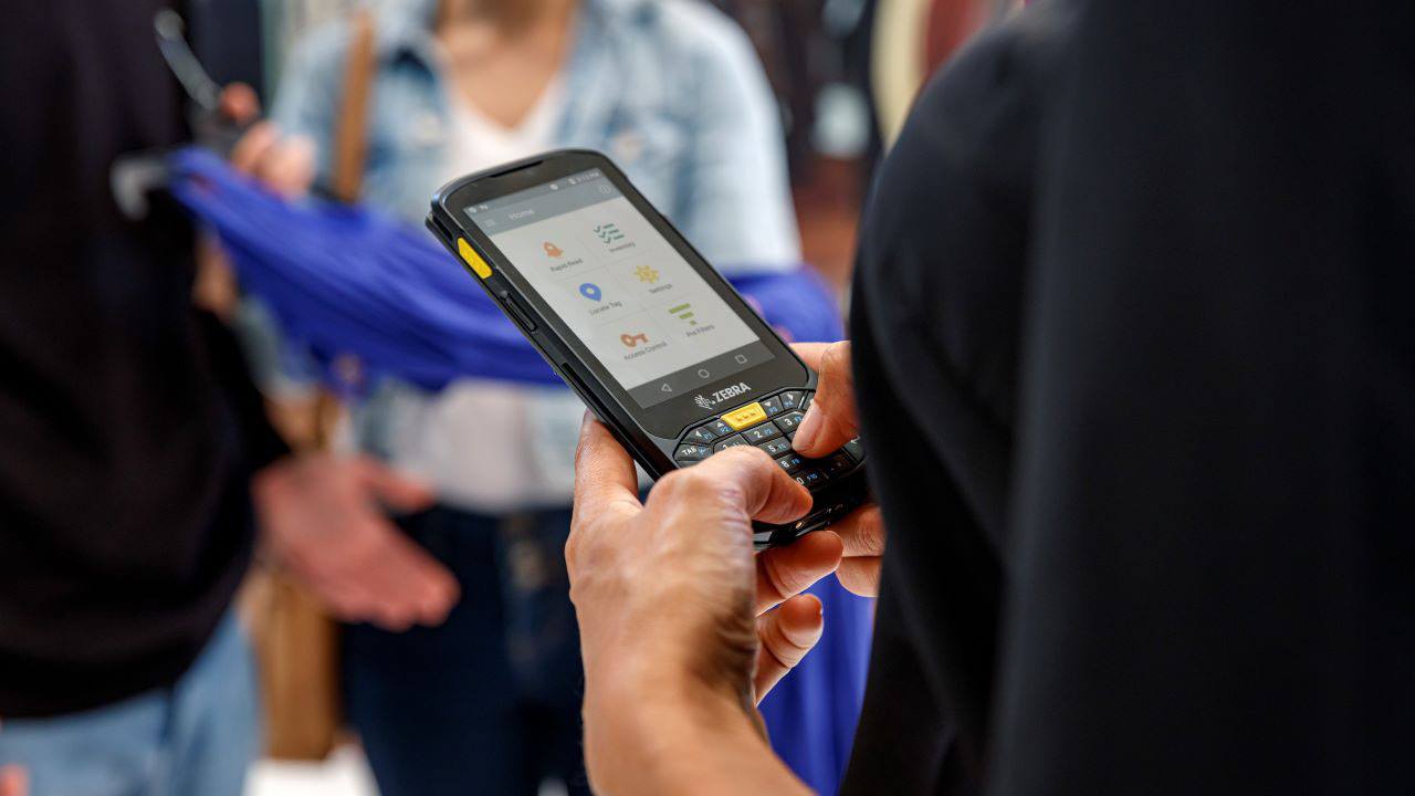 A department store associate uses a handheld mobile computer to look up something while keeping a safe distant from customers.