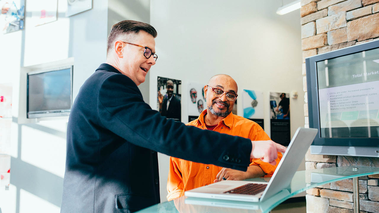 A technology consultant points to a laptop screen while meeting with a business manager