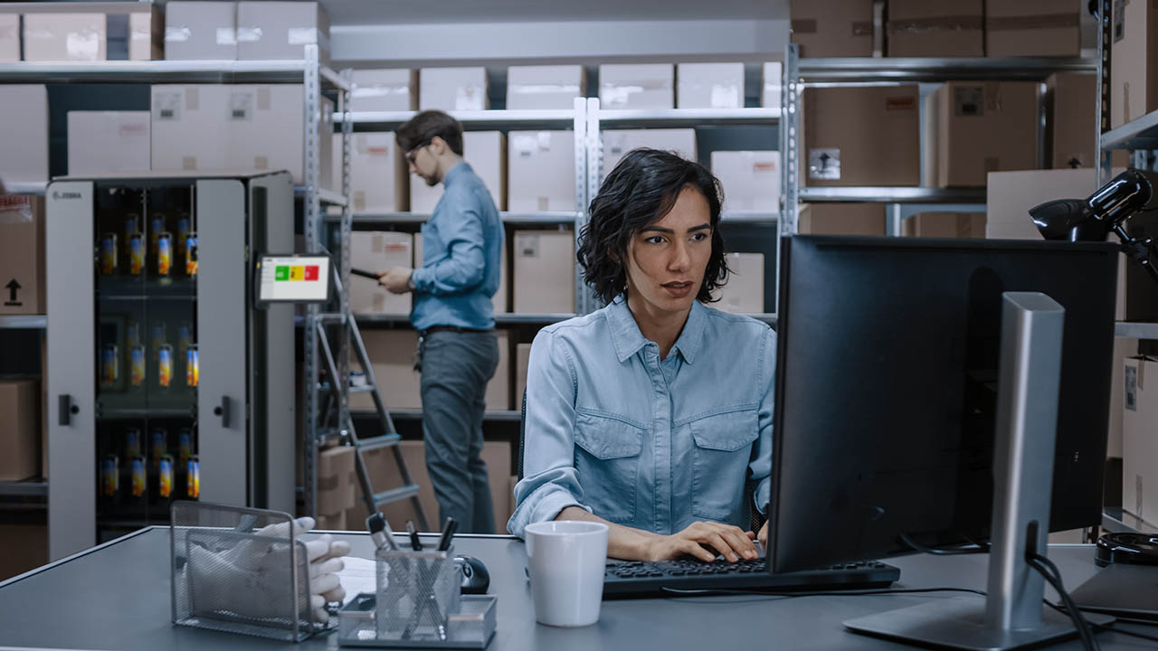 A consultant looks at a computer in the back of a warehouse while another takes inventory of mobile computers are stored