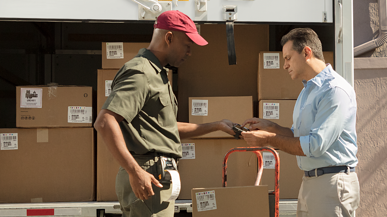 Delivery driver standing in front of a loaded truck, handing a package to a customer.