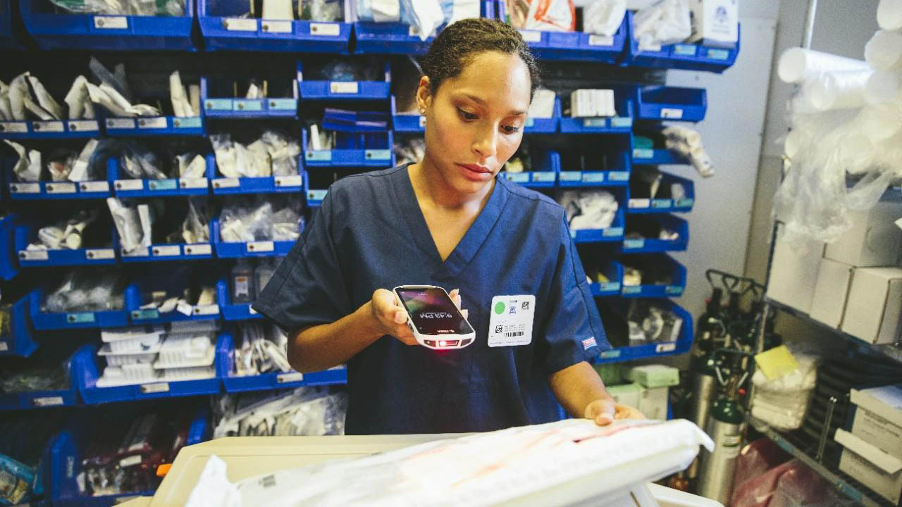 A nurse uses a clinical smartphone to scan a medical device package in a hospital stockroom.