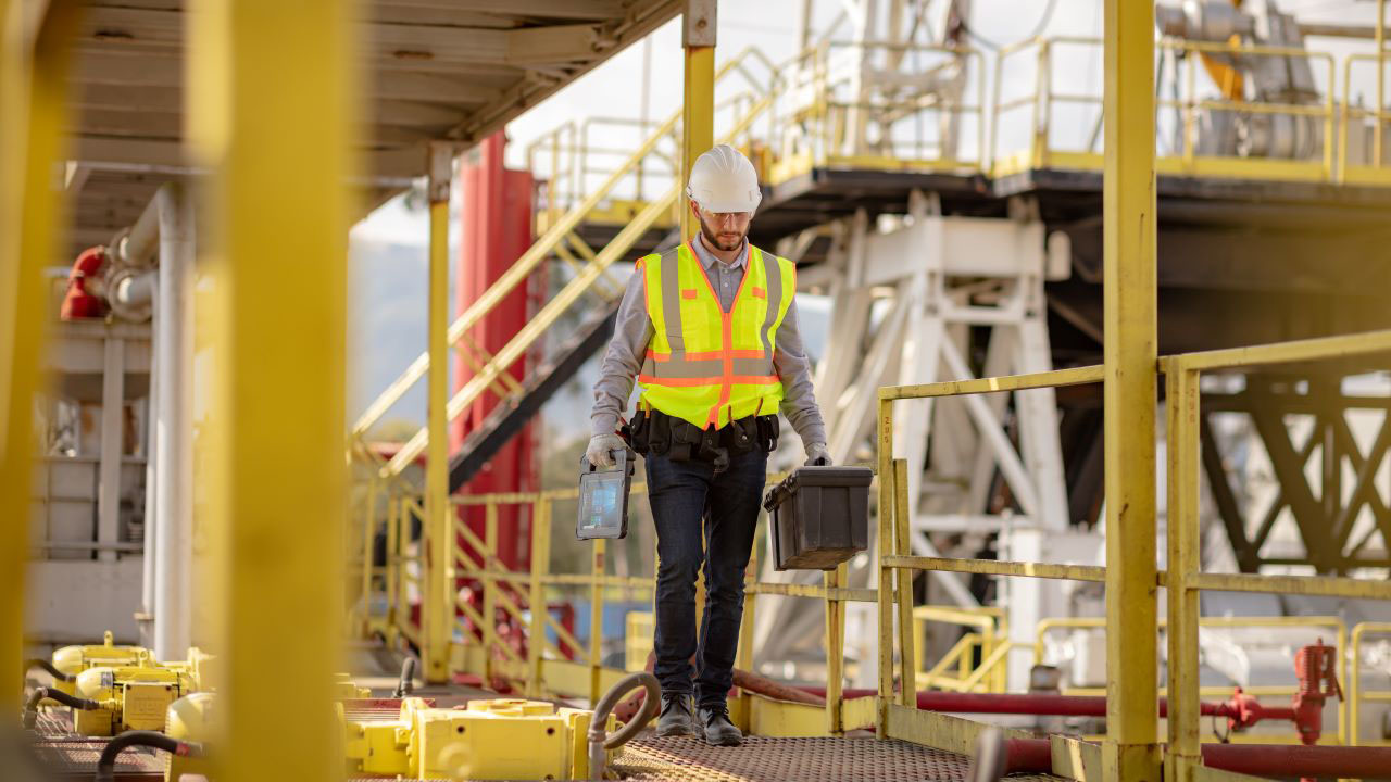 A utility field worker walks across a catwalk carrying a toolbox in one hand and a Zebra XSLATE L10 rugged tablet in the other.