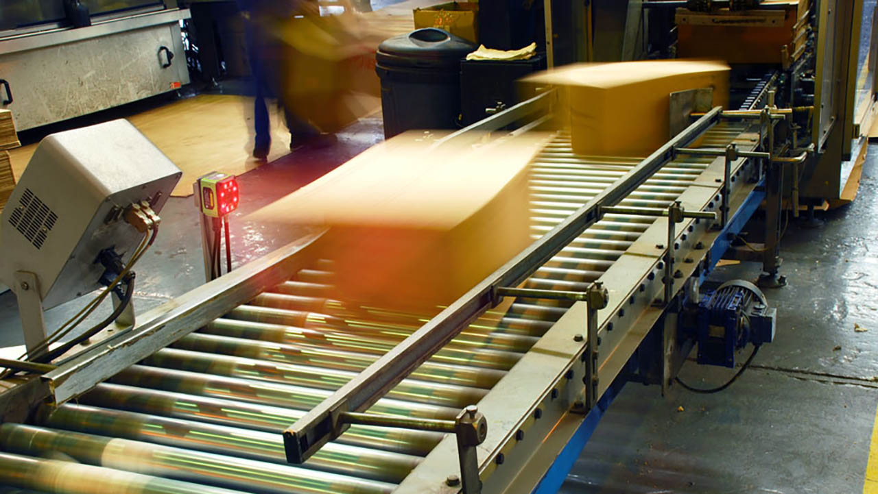 A fixed industrial scanner is used to scan barcoded boxes as they move down a conveyor belt in a warehouse.