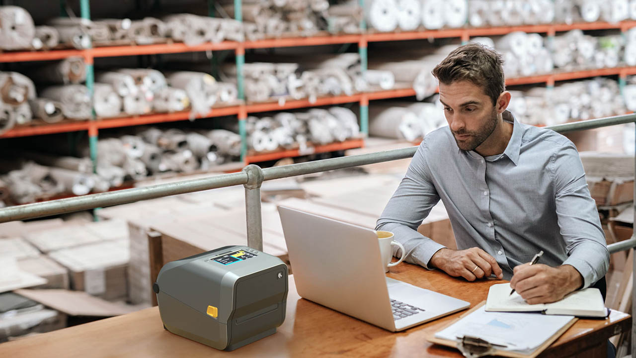 A man sitting at a desk in a warehouse takes notes while looking at a computer screen