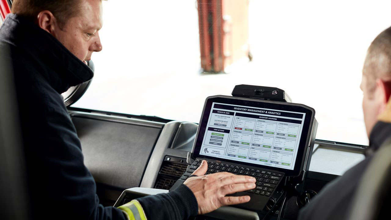 Two firefighters look at a rugged tablet mounted in the console of their truck.