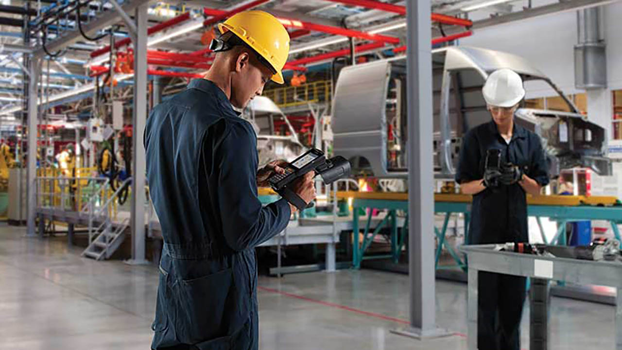 A manufacturing worker looks at his mobile computer while standing next to an auto assembly line.