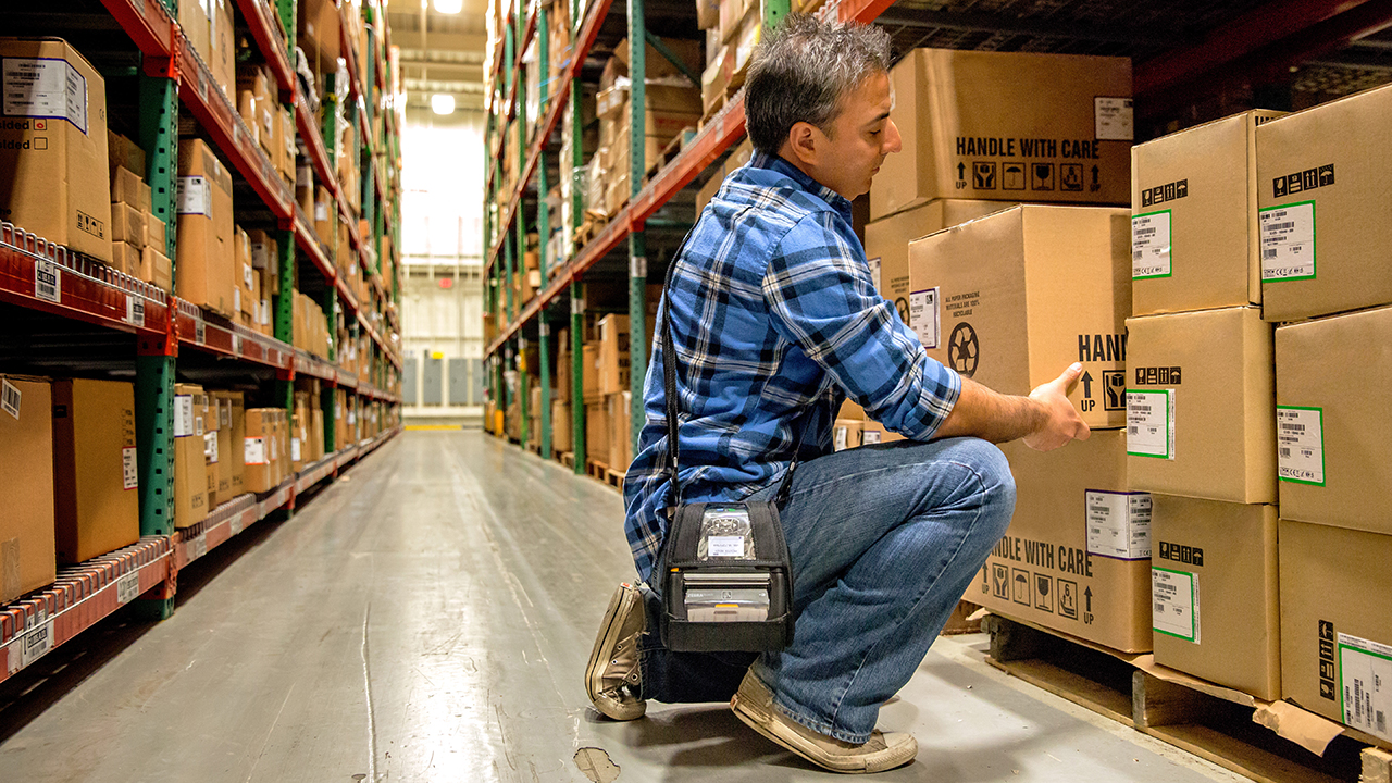 A warehouse worker wearing a mobile printer pulls a box off a shelf.