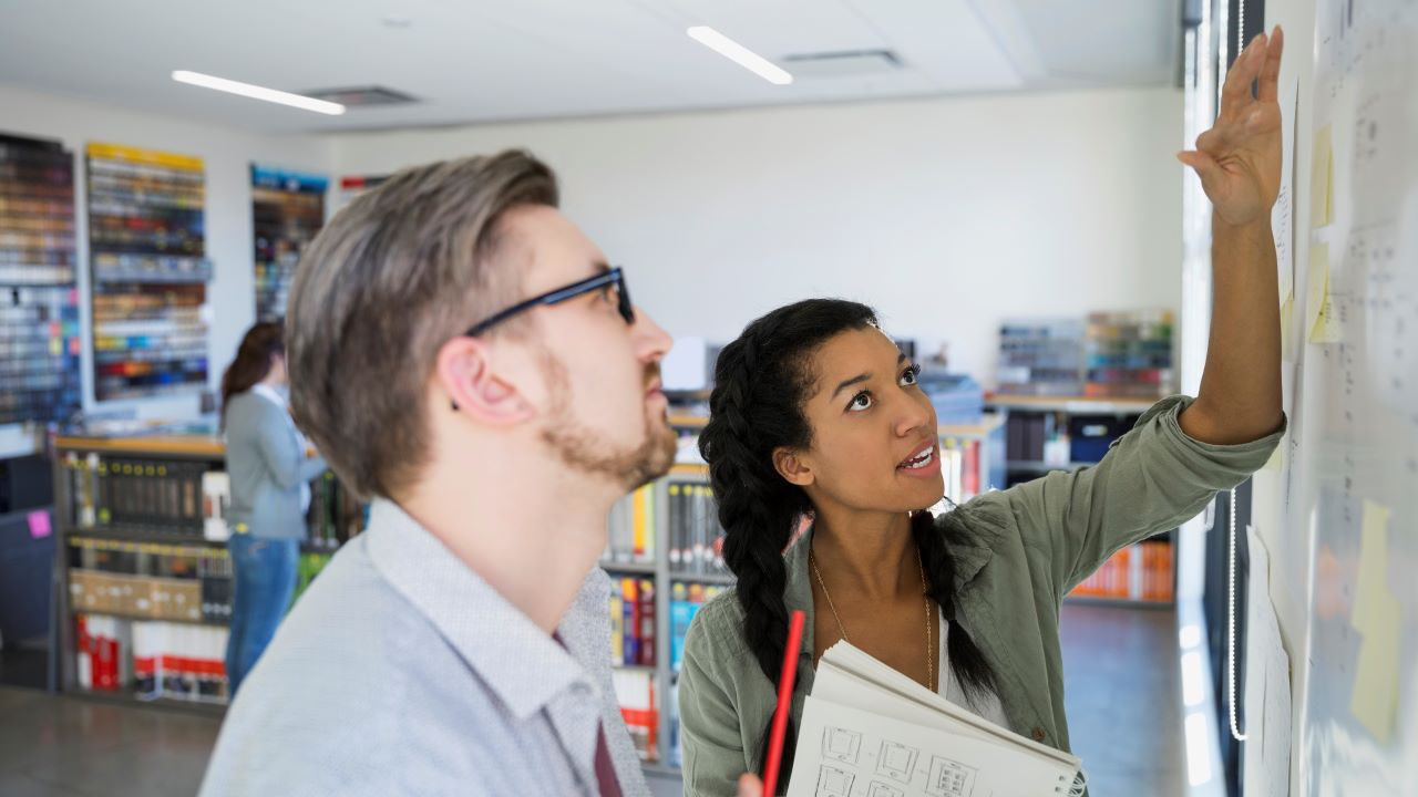 A man and woman review and discuss notes on a whiteboard.