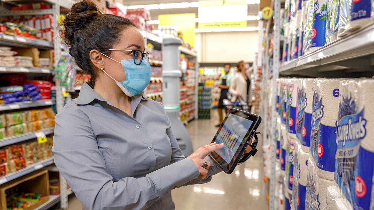 A grocery store manager reviews shelf inventory information on her rugged tablet in the toilet paper aisle