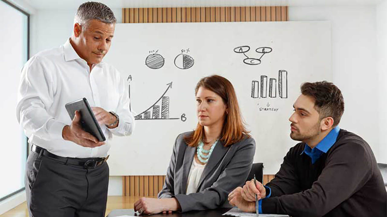 A professional services consultant shows a man and a woman something on a rugged tablet screen.