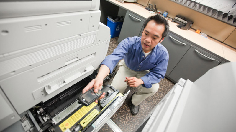 A worker uses an early model touch computer to inspect a piece of office equipment.