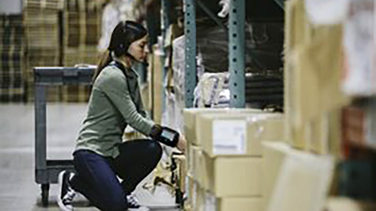 A female warehouse worker kneels down to grab a box from a low shelf