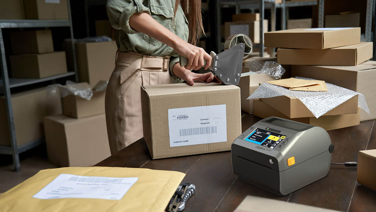 The Zebra ZD621D printer sits on the desk a woman is using to pack boxes