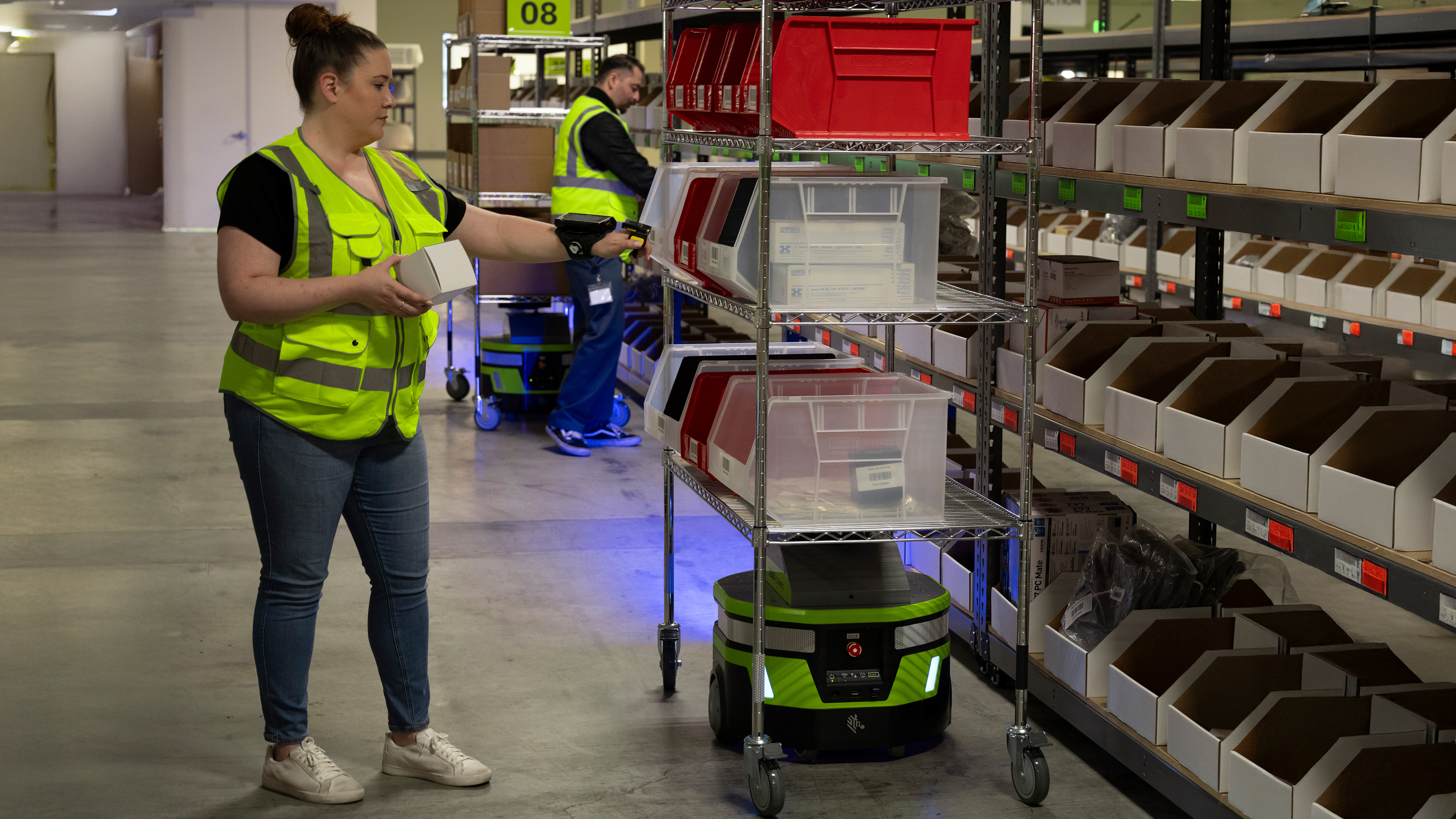 Warehouse staff holding a white box in front of the shelves and Zebra mobile robot