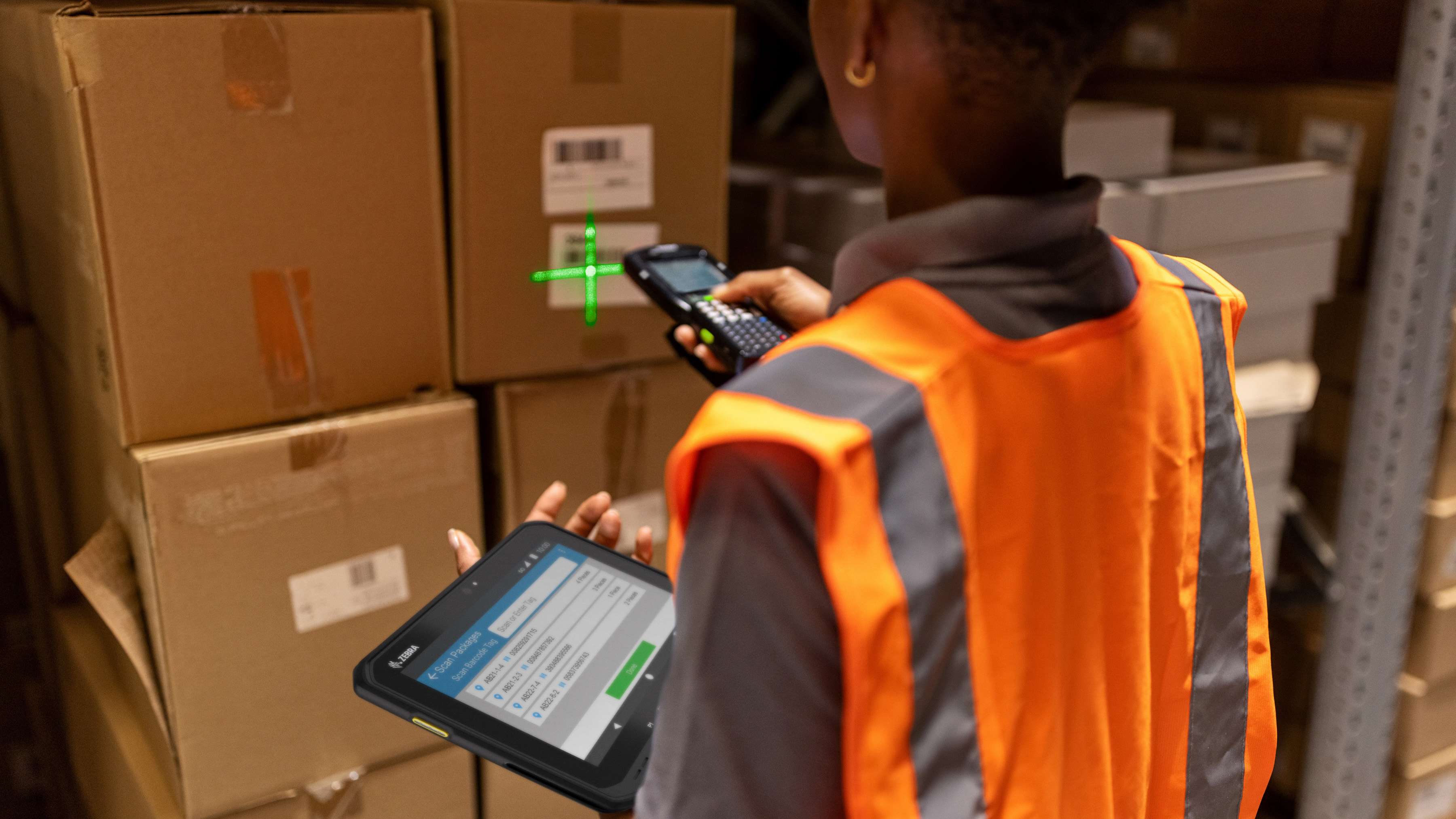 An employee using a Zebra handheld mobile computer and tablet to scan a box inside of a warehouse setting 