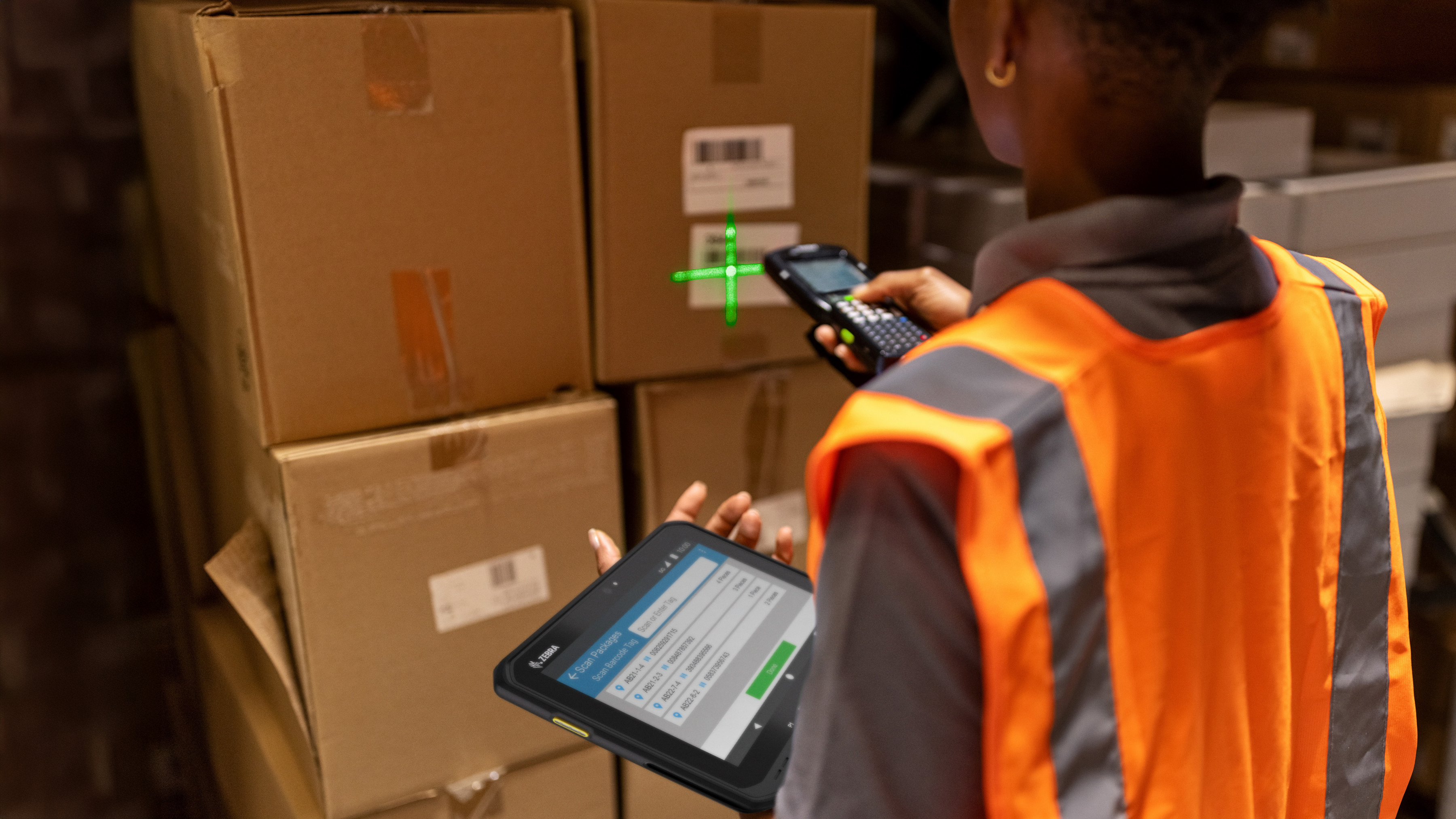 An employee using a Zebra handheld mobile computer and tablet to scan a box inside of a warehouse setting 