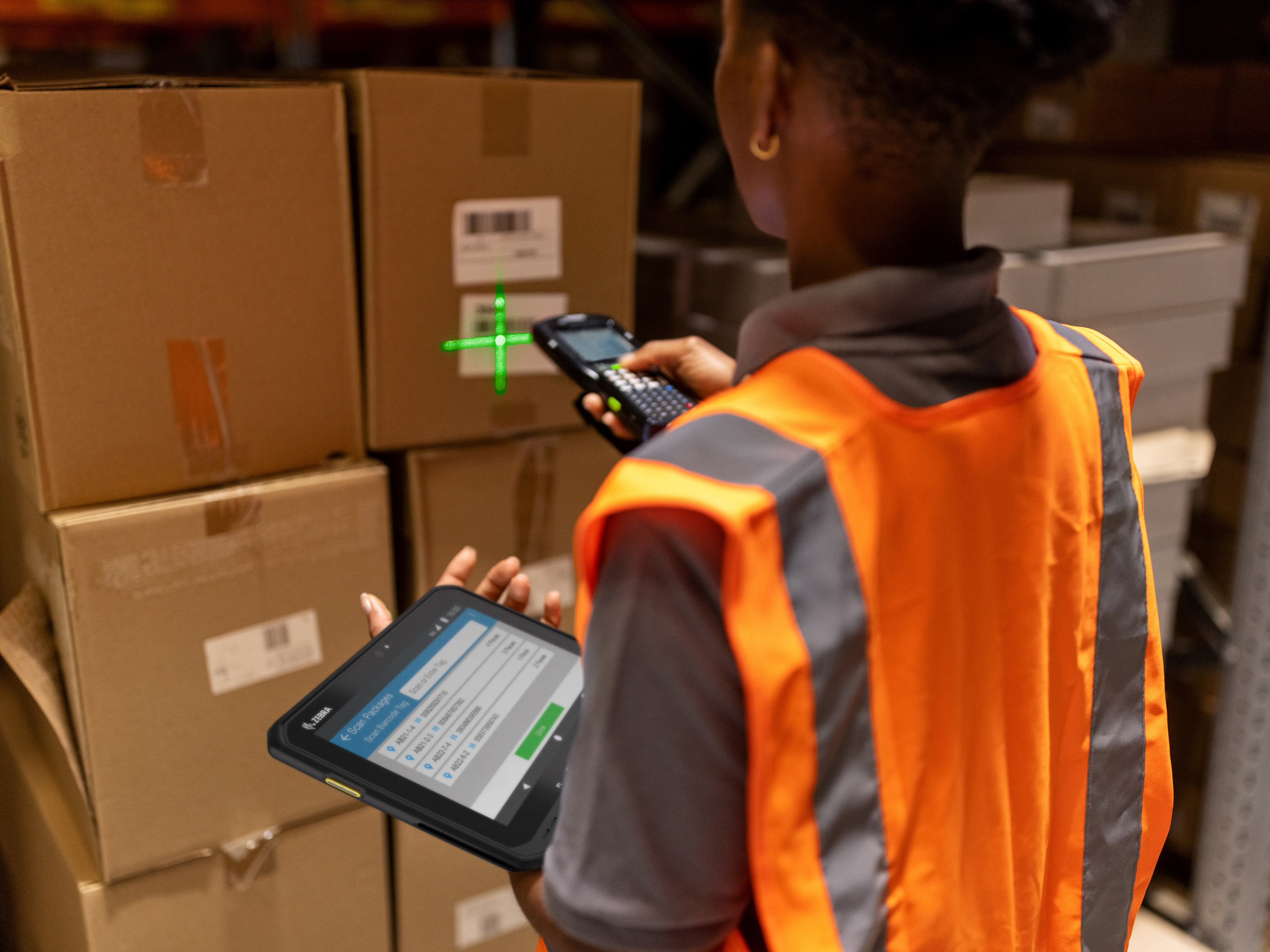 An employee using a Zebra handheld mobile computer and tablet to scan a box inside of a warehouse setting 