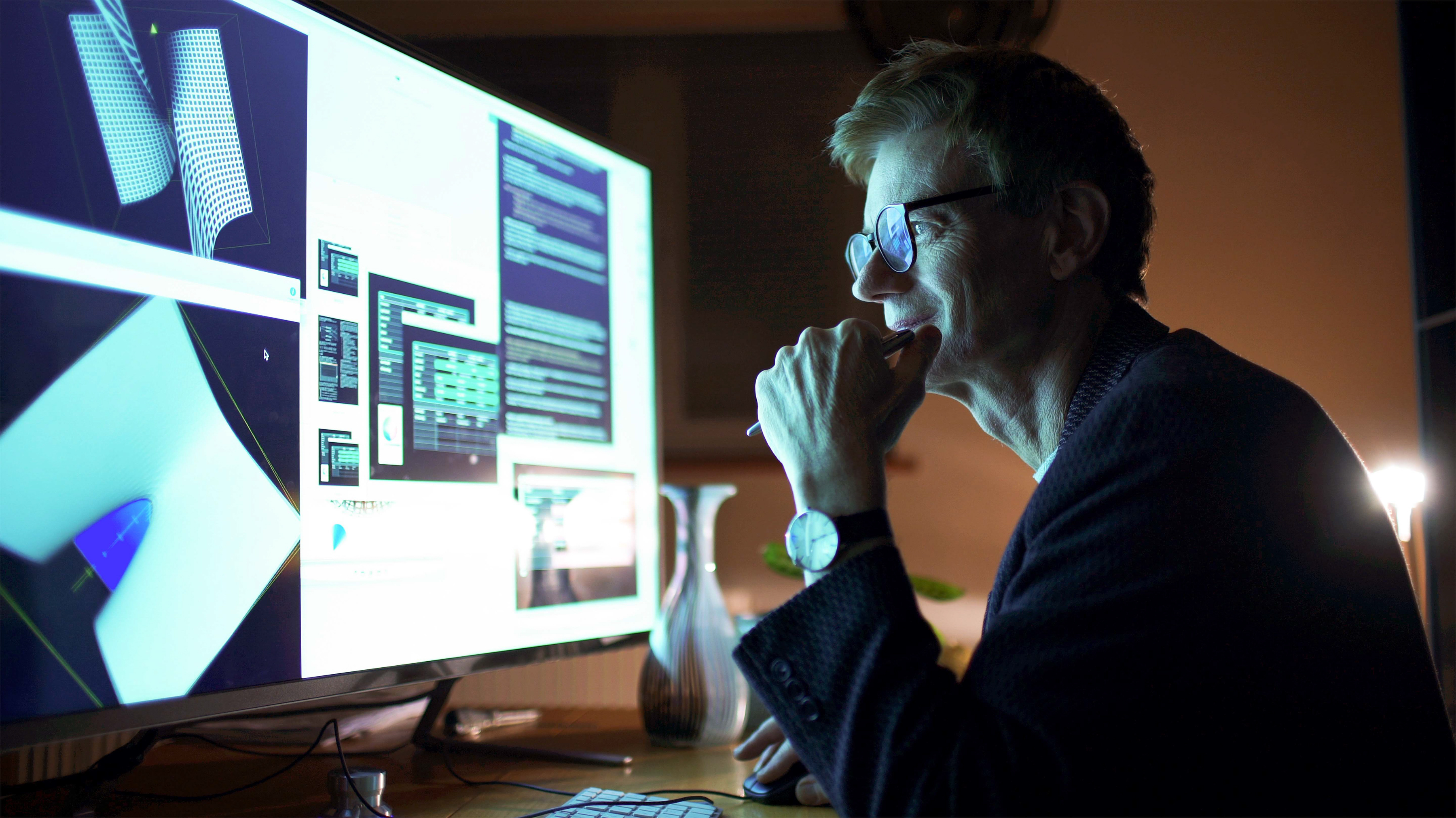 An employee works late into the night at a desktop computer, where a large monitor displays a various image models and code.