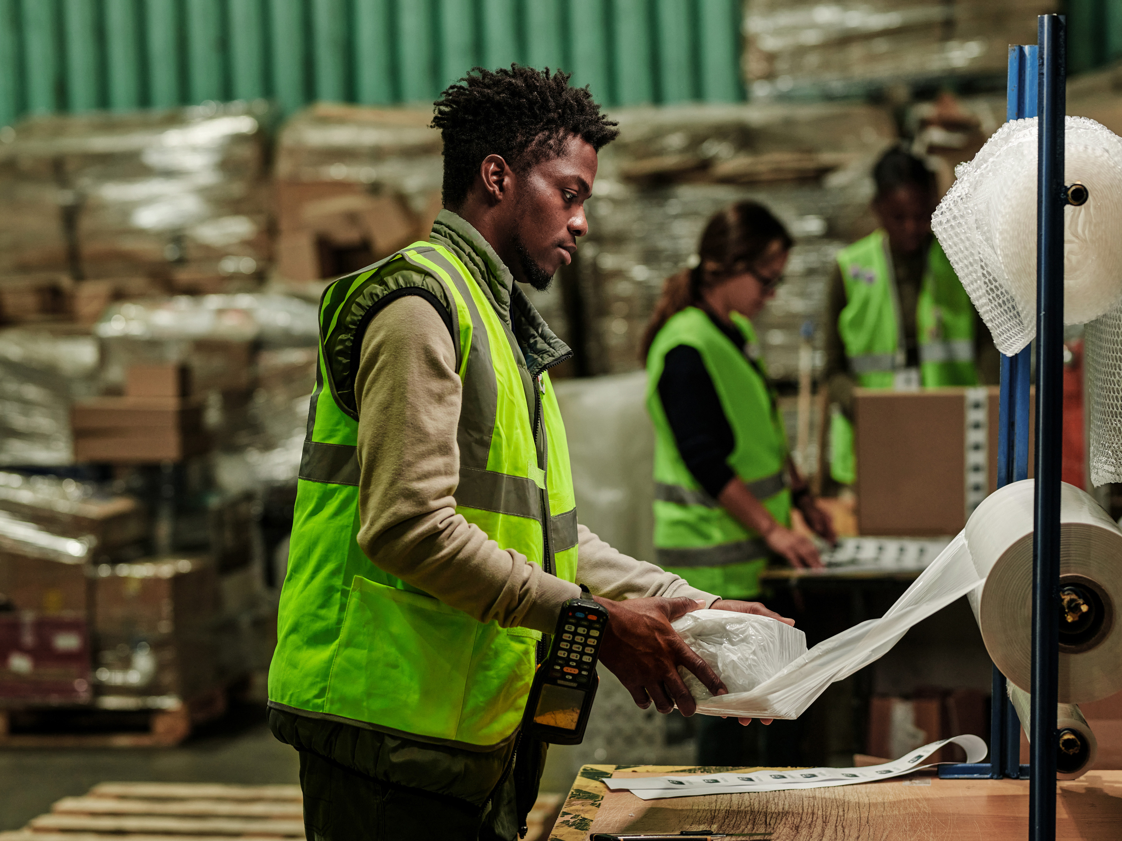 A worker in a warehouse has a handheld computer securely hanging from his wrist while packaging items using protective wrap for shipment.