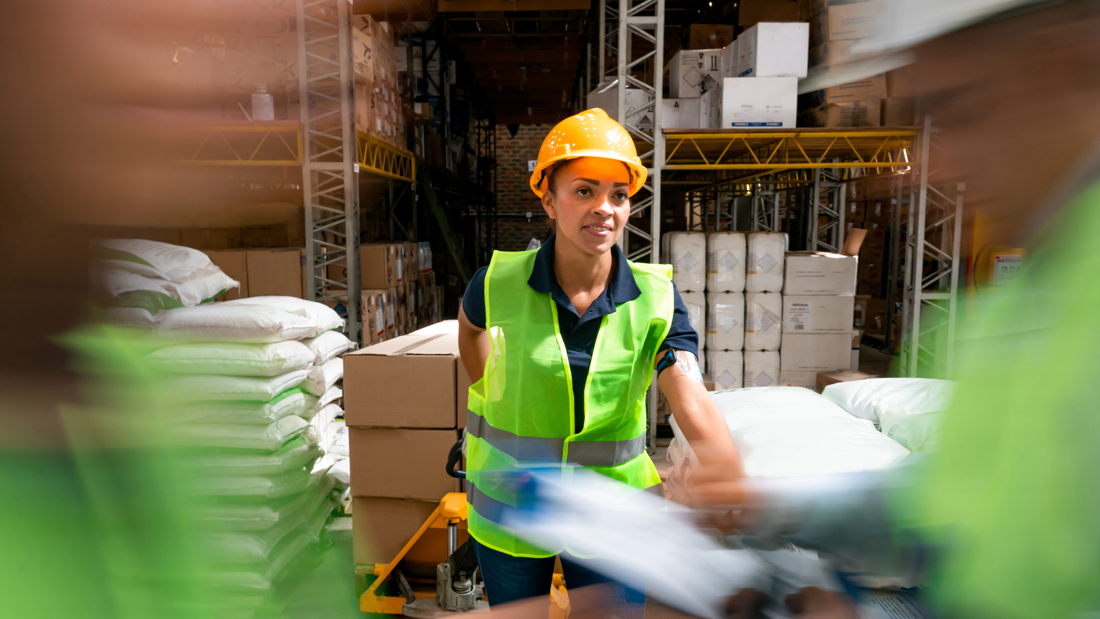 A worker in a busy distribution warehouse wearing a hard hat and hi-vis safety vest moves boxes using a forklift.